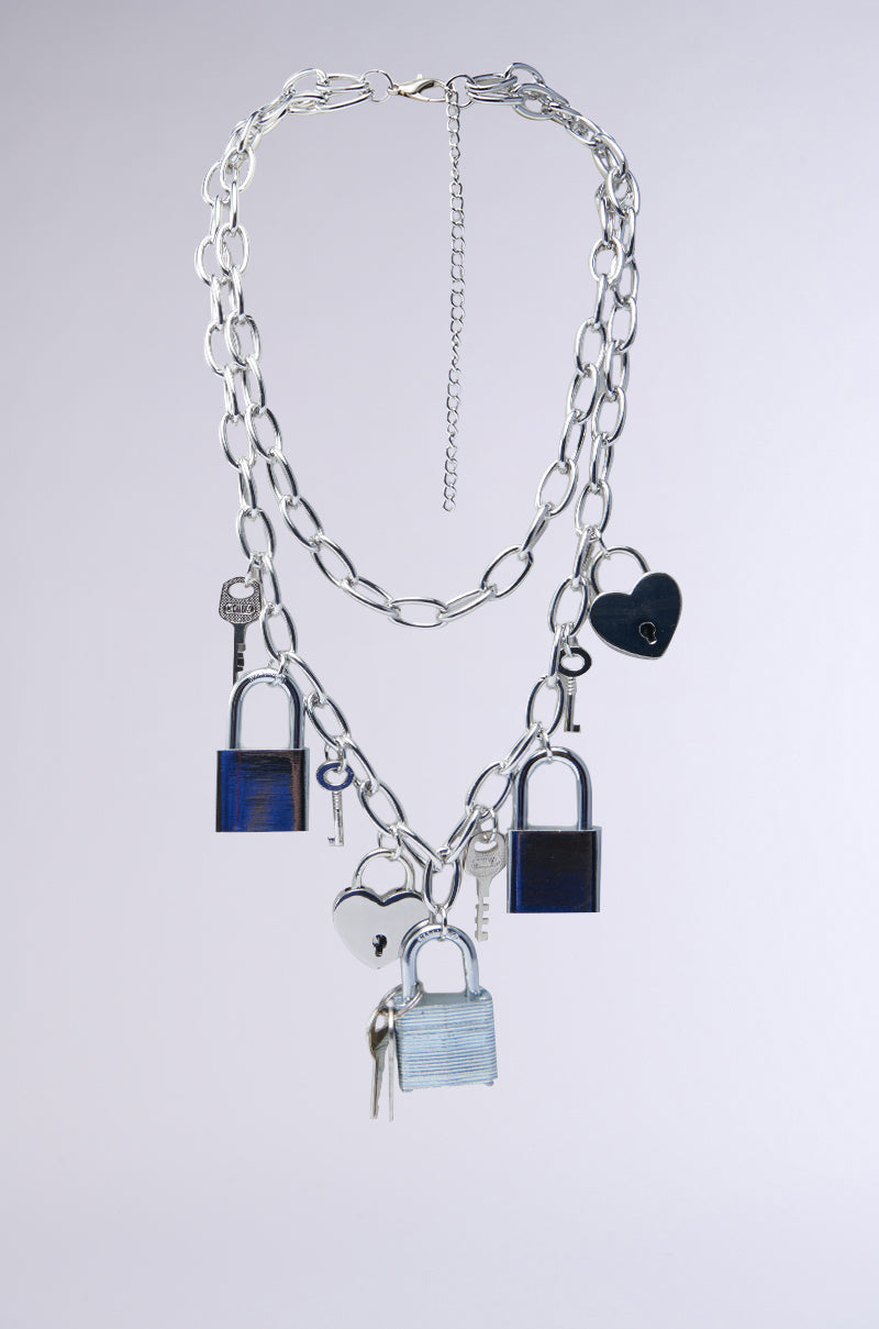 Meet the HEART ON LOCK CHAIN NECKLACE, a silver chain featuring adorable heart-shaped and sleek square locks and keys. This padlock accessory shines brilliantly against its light gray backdrop.