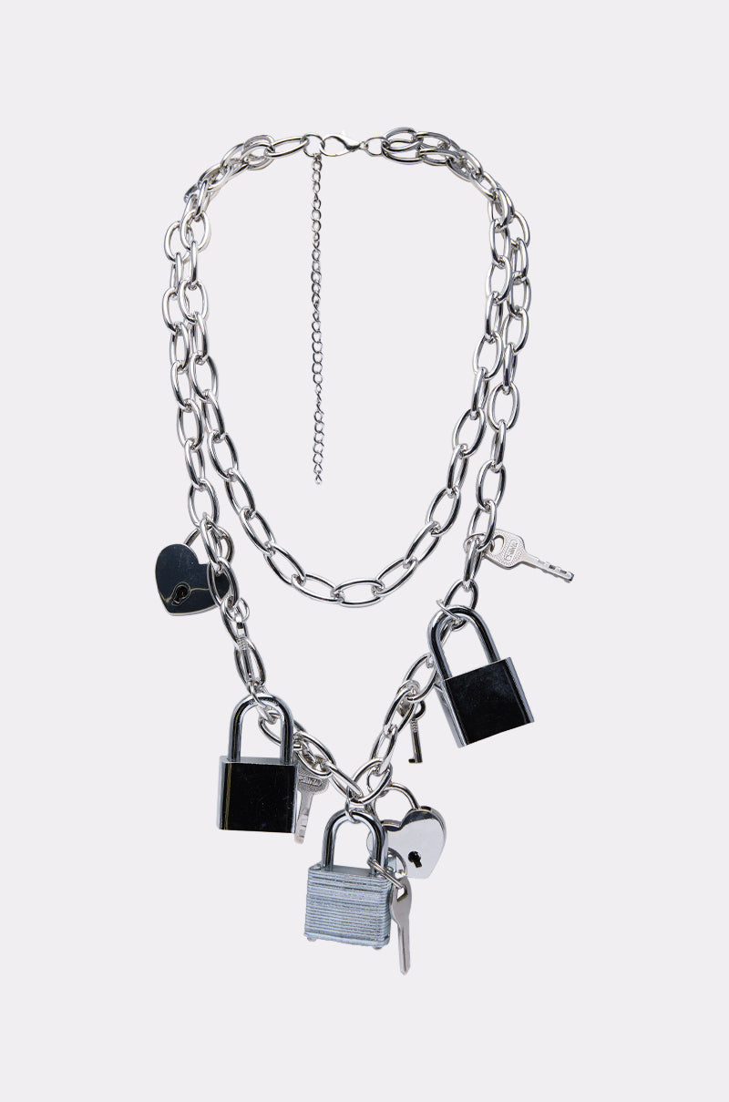 The HEART ON LOCK CHAIN NECKLACE is a silver chain with large links, featuring three padlocks and three keys attached at intervals, styled as a padlock necklace and shown on a plain light gray background. 0