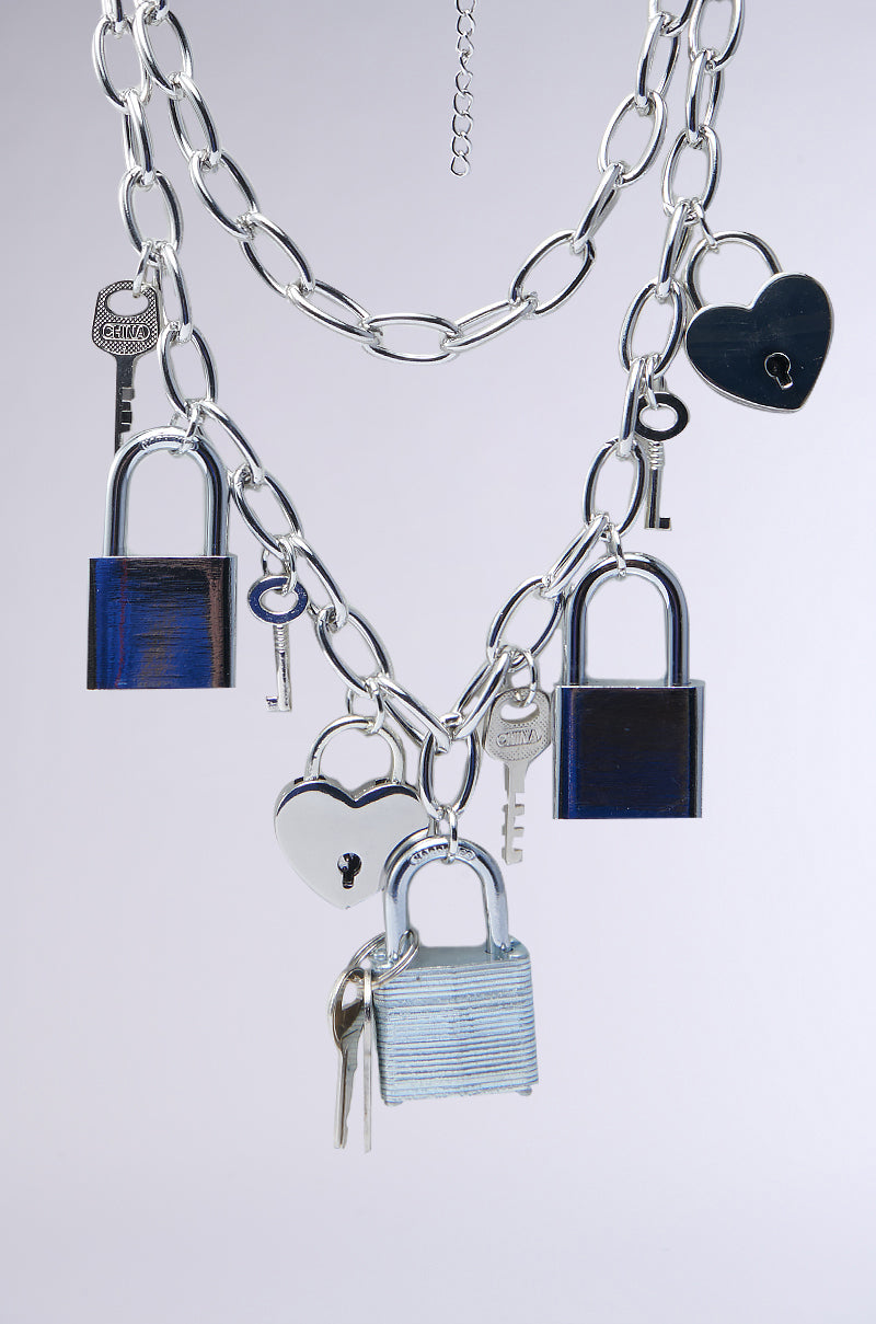 The HEART ON LOCK CHAIN NECKLACE features a silver chain with heart-shaped lock and key pendants, creating an elegantly intertwined look against a plain backdrop.