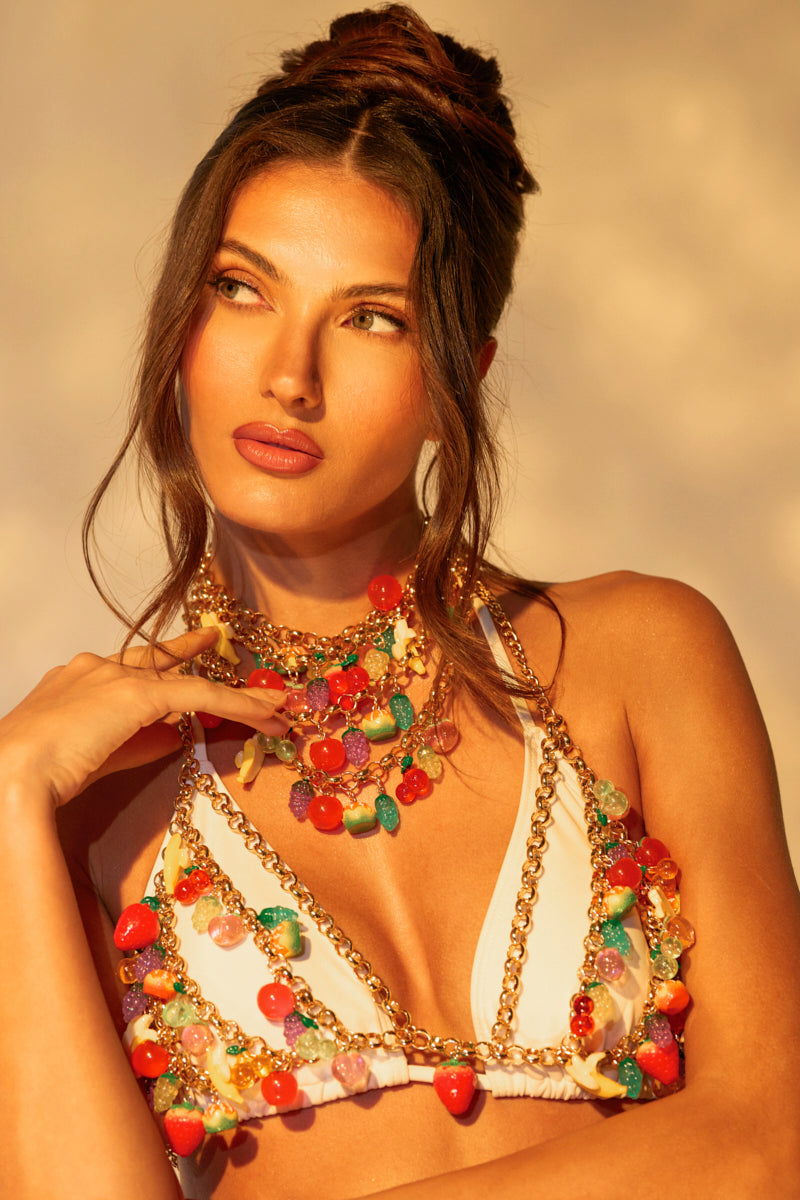 A woman with long dark hair tied up wears a vibrant FEELIN FRUITY NECKLACE featuring colorful beads and fruit-shaped charms, posing against a softly lit background. She gazes thoughtfully to the side, exuding confidence and poise.