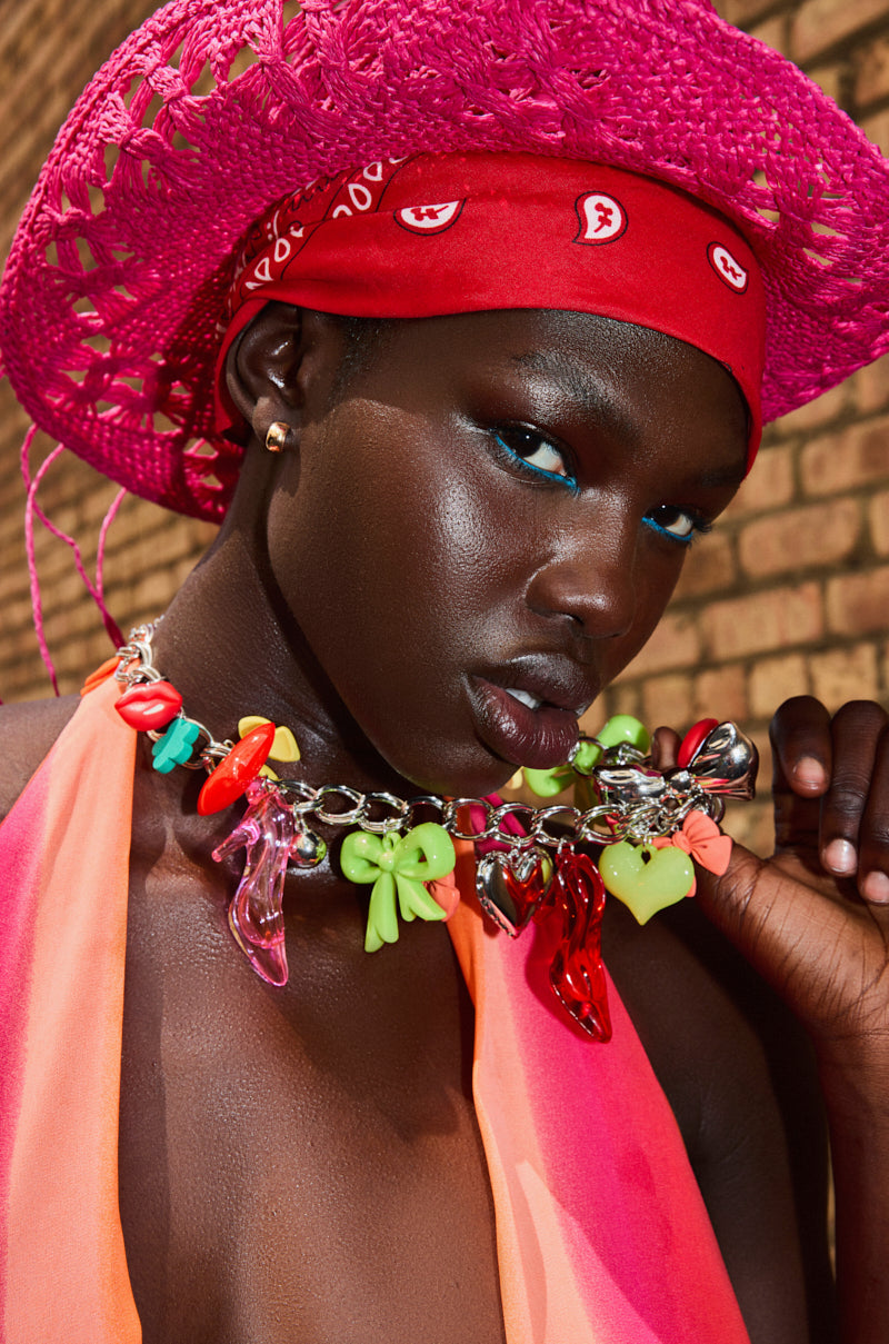 A woman in a bright pink hat, red bandana, and the CHARM LIFE NECKLACE with colorful charms poses confidently. She wears bold makeup, gold stud earrings, a pink and orange halter top, all set against a brick wall background. 0