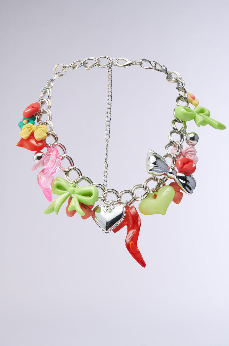The CHARM LIFE NECKLACE is a chunky chain adorned with colorful charms—bows, hearts, a chili pepper, and a metallic heart—creating a bold statement piece, shown on a light gray background. 1