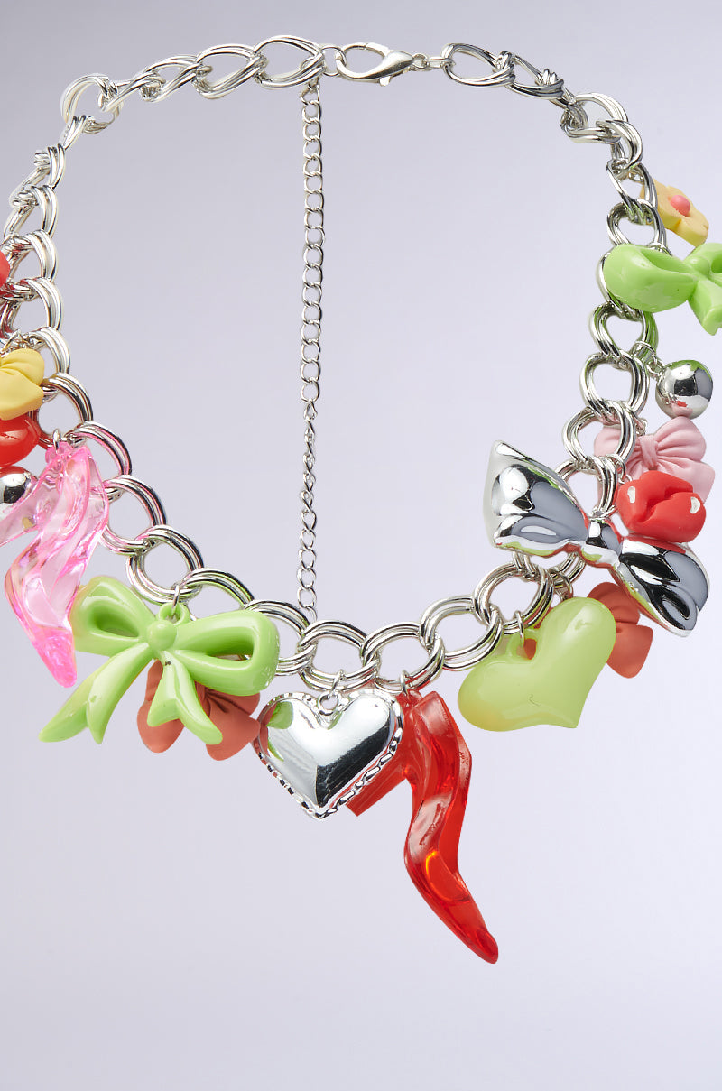 The CHARM LIFE NECKLACE is a bold statement piece featuring a chunky chain with colorful charms—hearts, bows, high heels, and abstract shapes in red, green, pink, yellow, and orange—shown against a light background. 4