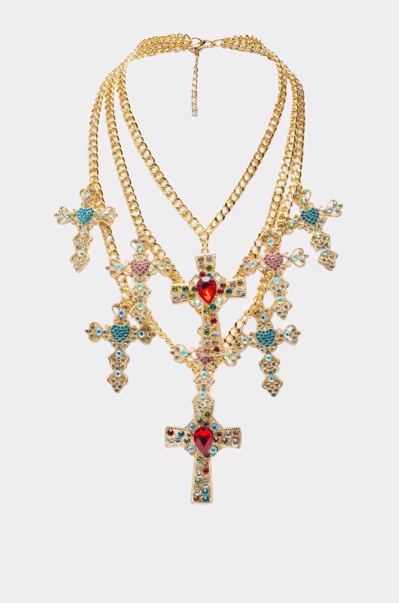 The IN MY FEELINGS NECKLACE is a multi-layered gold chain statement piece with ornate cross pendants, each embellished with colorful gemstones and a bold red heart-shaped gem at the center of the largest cross.