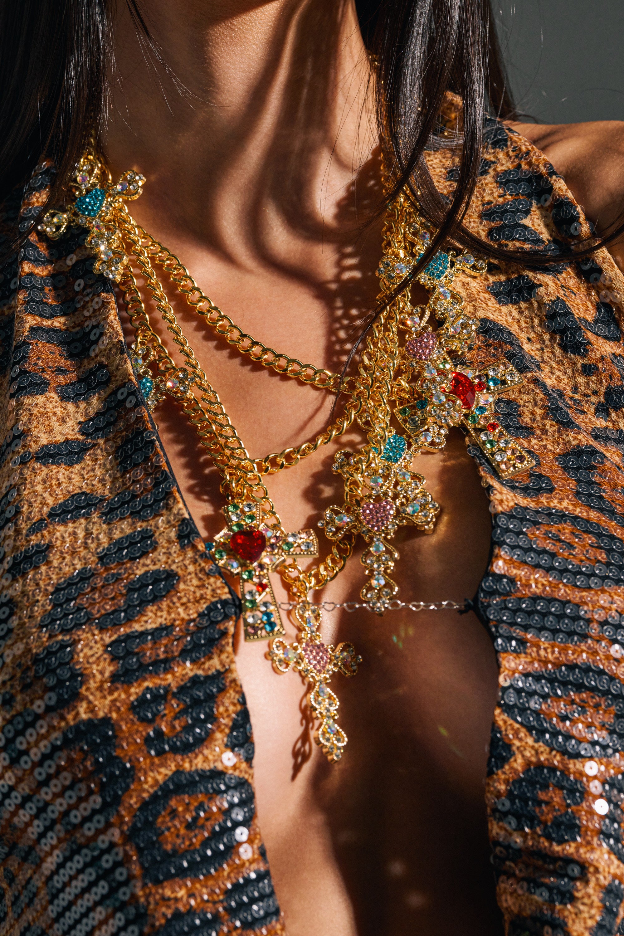 A woman wears the IN MY FEELINGS NECKLACE with ornate jeweled cross pendants layered over a sequined leopard print deep V top. Sunlight highlights her skin and the shimmering jewelry.