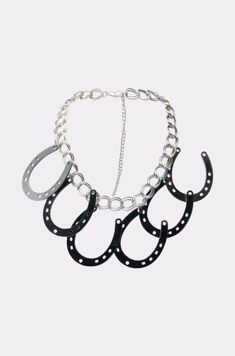The TEST MY LUCK HORSESHOE NECKLACE features five evenly spaced horseshoe-shaped pendants in silver to black on a chunky chain, making a bold statement against a plain white background. 0
