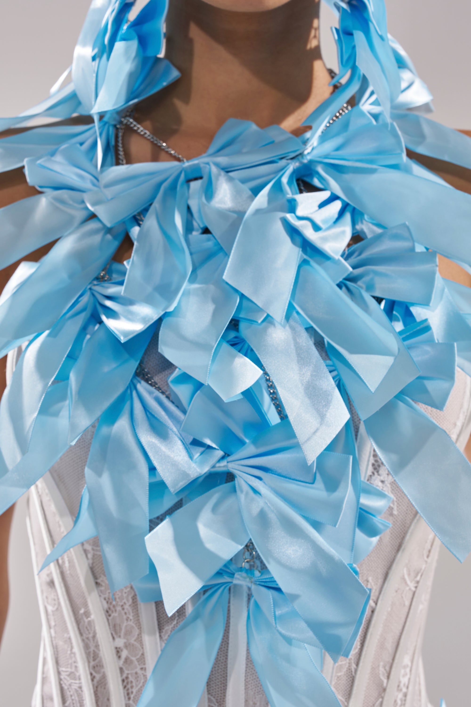 A person models the FEMME BOW STATEMENT NECKLACE, featuring large, shiny light blue satin bows in a sculptural, layered design that covers the chest and shoulders for a bold, feminine jewelry look. 1