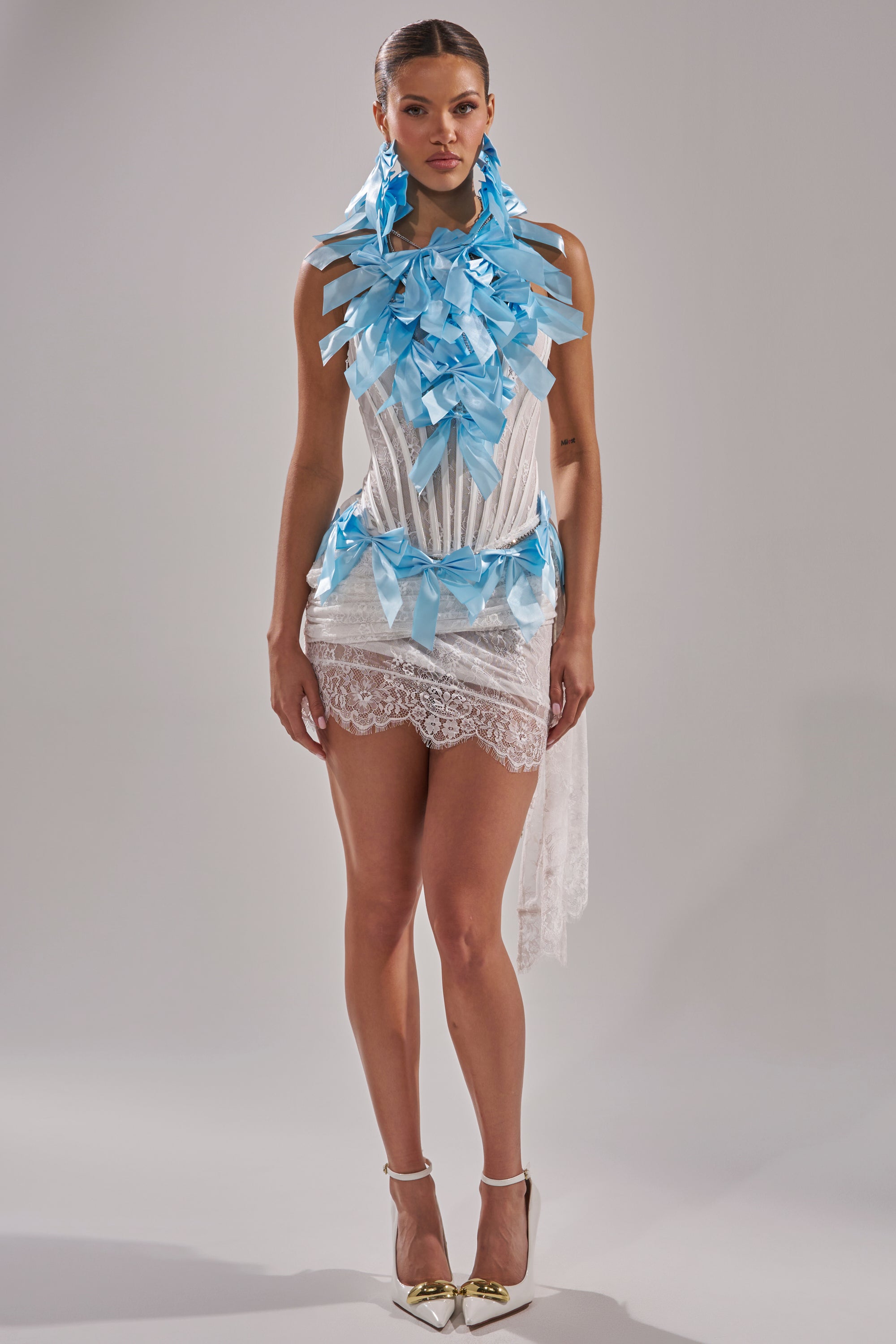 A woman stands facing forward in a short, sheer white lace dress, accentuated by the FEMME BOW STATEMENT NECKLACE featuring large, layered blue ribbons and paired with gold heels against a plain background.
