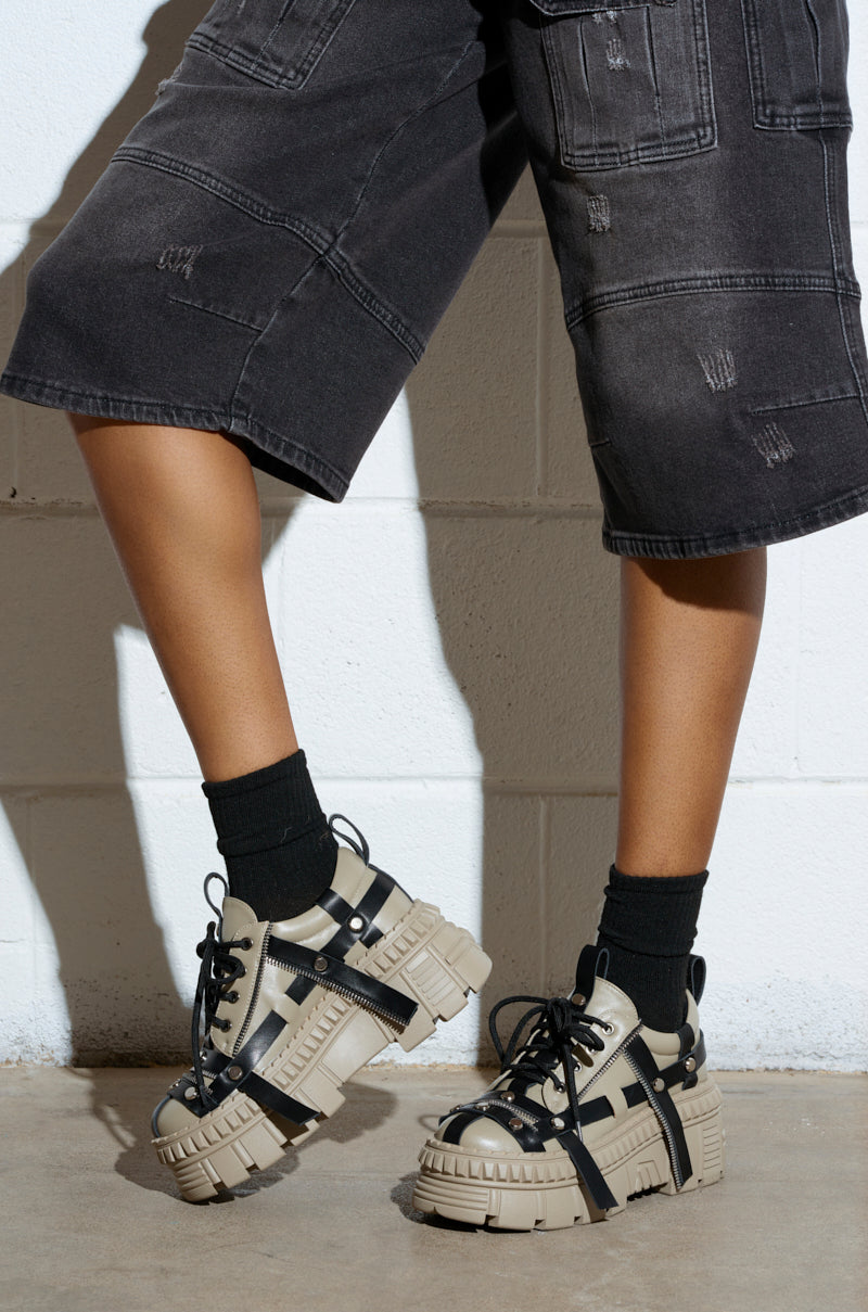 Wearing knee-length black denim shorts and AZALEA WANG BEAGLE KHAKI FLAT SNEAKERS with contrasting black lace, a person showcases a cool style with black socks against a white brick wall on a concrete floor.