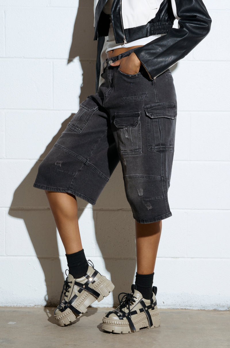 A person in a black and white cropped jacket and distressed black cargo shorts stands against a white wall, wearing black socks with AZALEA WANG BEAGLE KHAKI FLAT SNEAKERS, one hand casually in a pocket, with their face unseen.