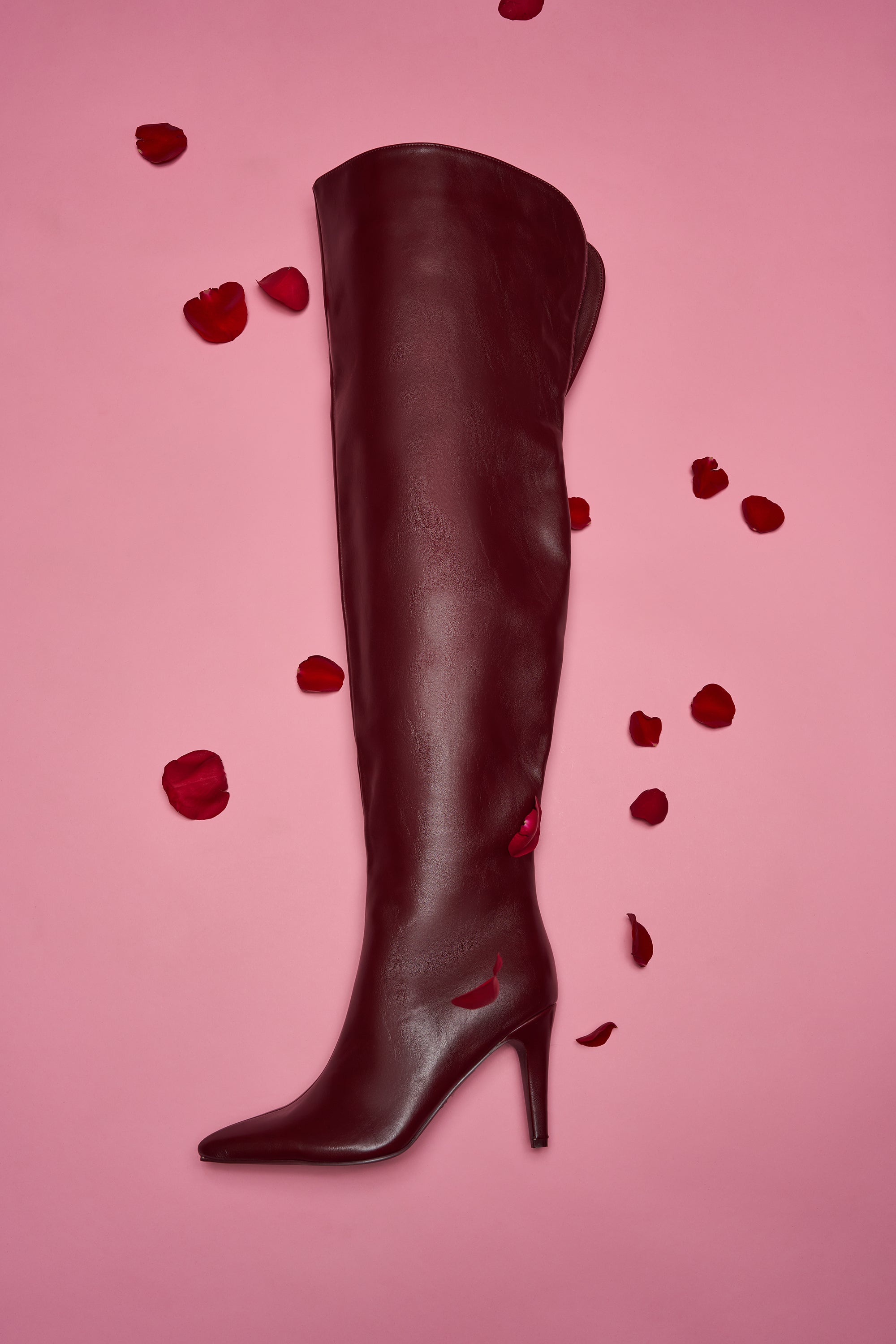 The AZALEA WANG BEAUCOUP BURGUNDY OVER THE KNEE BOOT, featuring a stiletto heel, is displayed on a pink background with red rose petals scattered around. 0