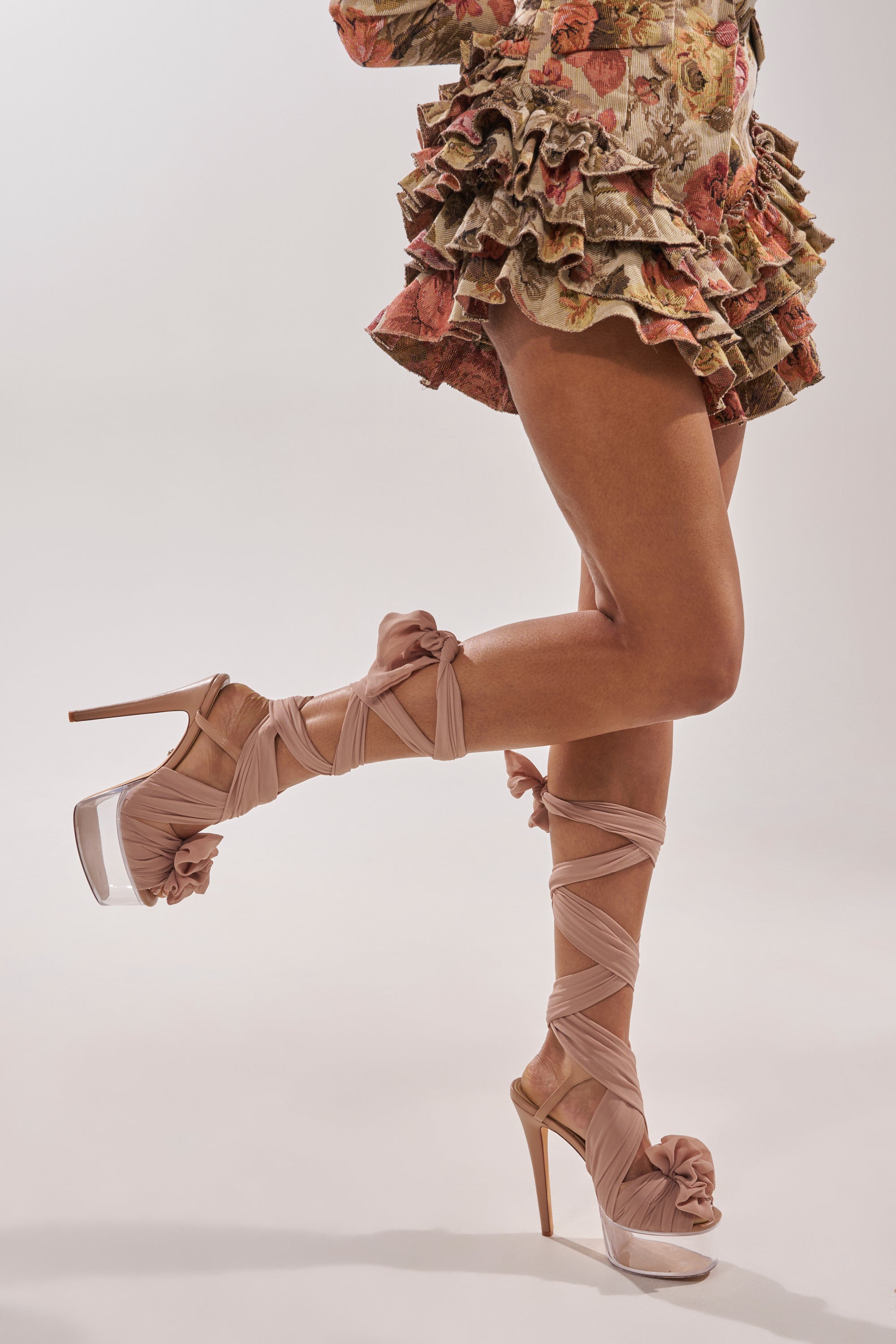 A person wearing floral ruffled shorts and AKIRA GOLD BEAUDEN TAN PLATFORM TIE SANDAL with square toe heel detailing, ribbons wrapped around crossed legs, stands against a plain light background. 0