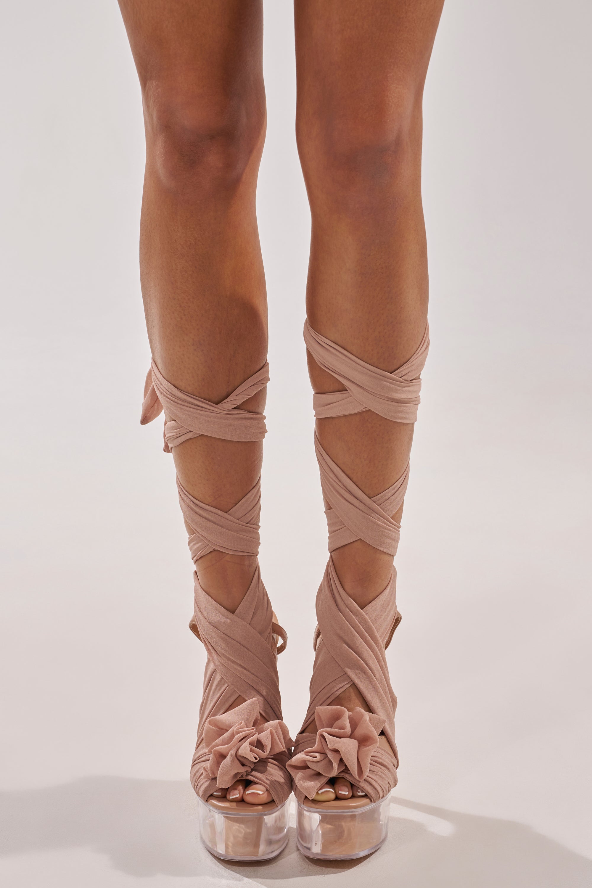 A person wears the AKIRA GOLD BEAUDEN TAN PLATFORM TIE SANDAL, featuring clear platform heels and nude, ruffled fabric wrapped in a crisscross pattern up the calves, standing against a plain light background. 1
