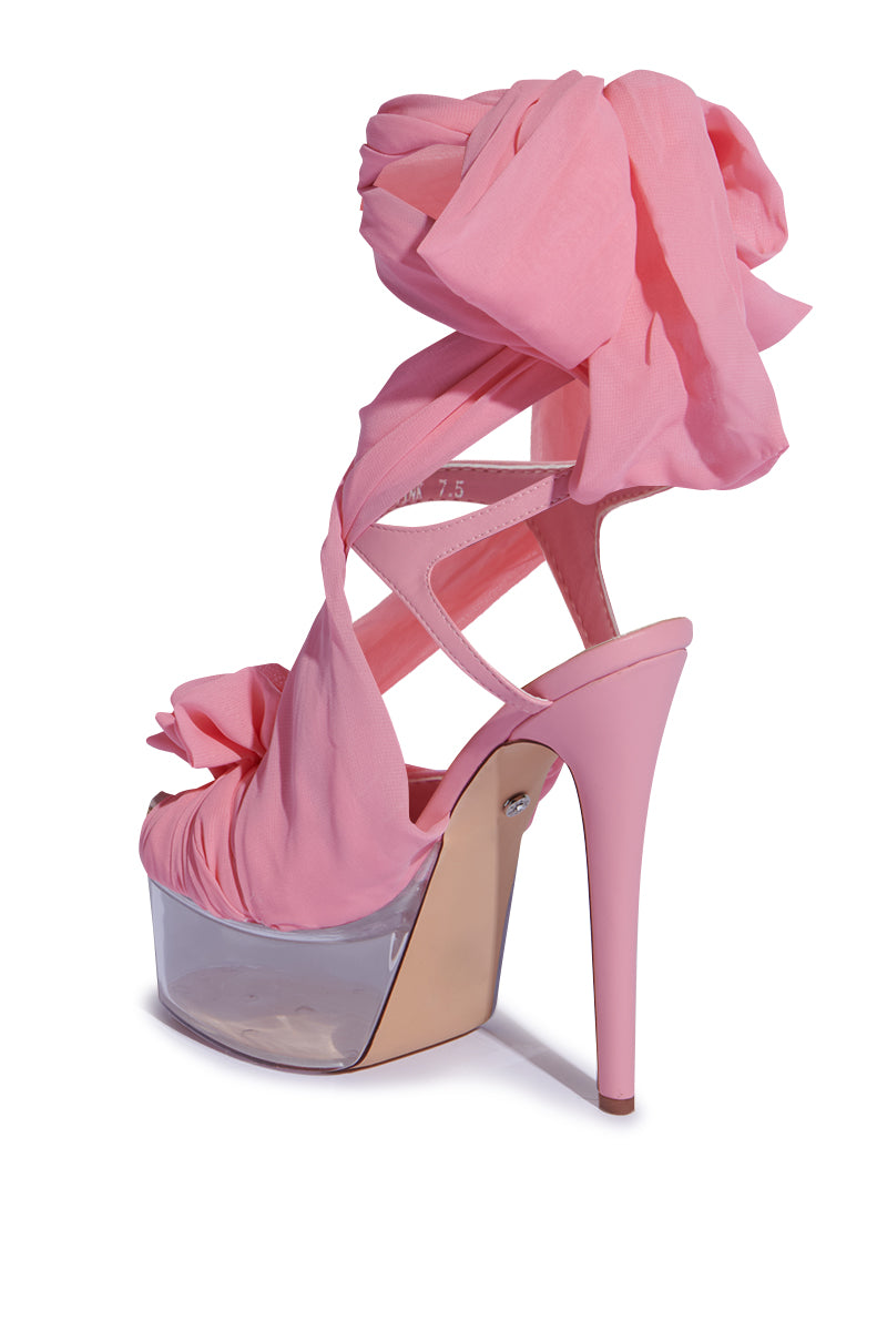 The AZALEA WANG BEAUDEN PINK STILETTO SANDAL features a clear platform sole and light pink stiletto with crisscrossing wraparound pink fabric ties, giving it an elegant bow-like appearance.