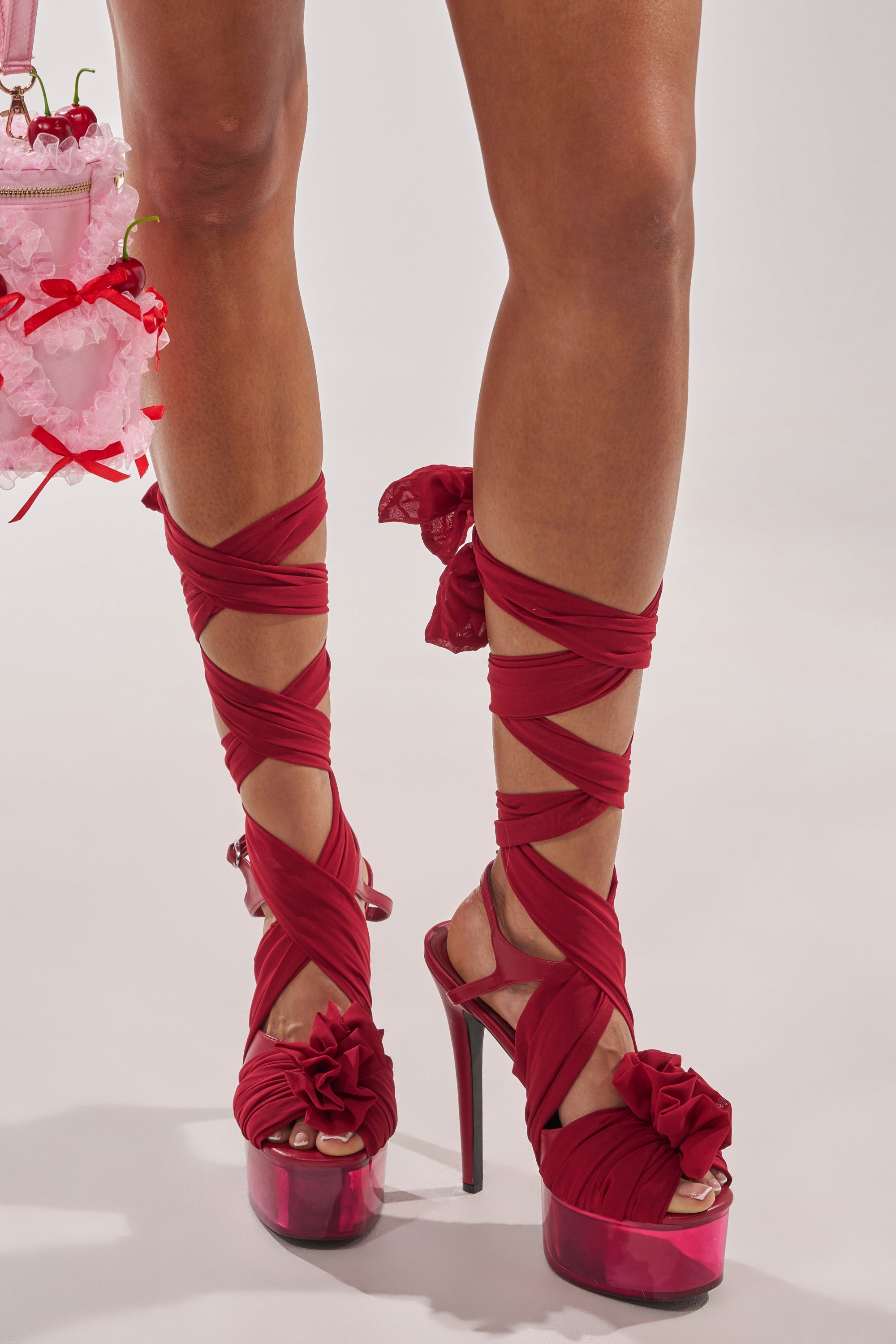 Someone is wearing the AKIRA GOLD BEAUDEN RED PLATFORM TIE SANDAL, featuring red fabric straps that crisscross and tie with bows up the legs. They are also holding a pink frilly accessory adorned with red ribbons. 0