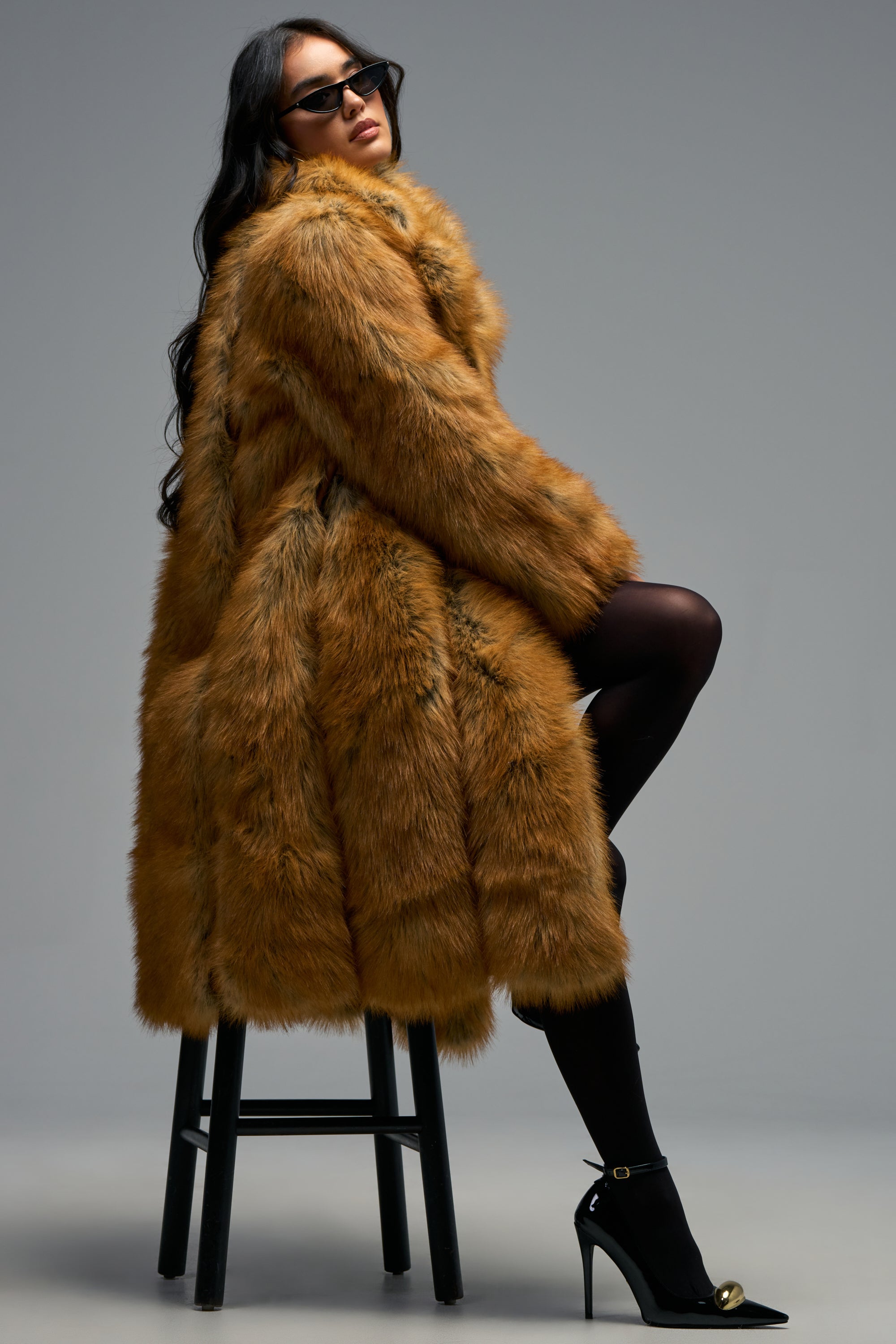 A woman sits stylishly on a stool against a plain gray background, embodying the elegance of winter layering in the BEAUMONT MAXI FAUX FUR coat, paired with black tights, high heels, and sunglasses.
