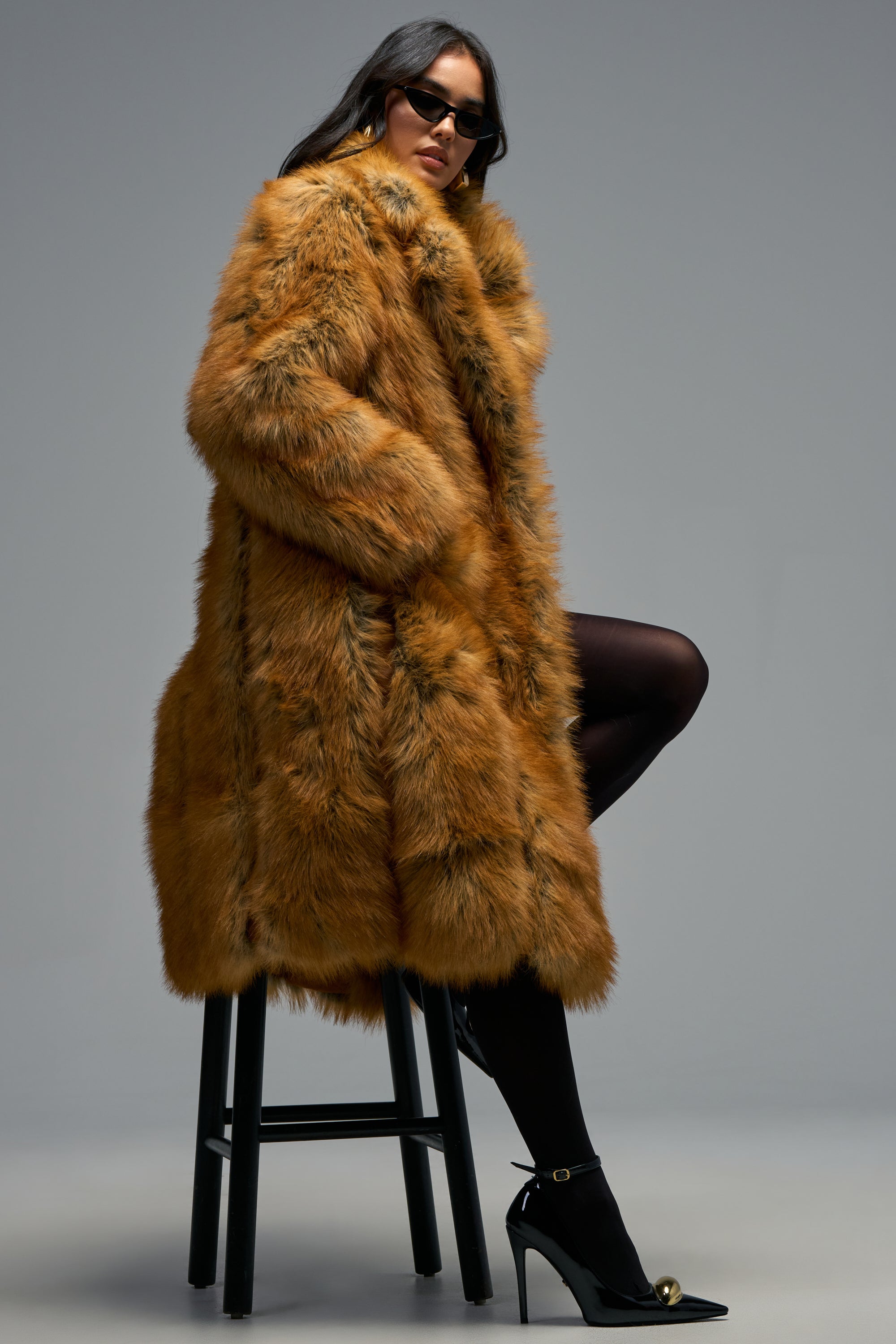 Wearing the BEAUMONT MAXI FAUX FUR coat, a person in sunglasses sits on a black stool, styled with black tights and high-heeled shoes against a plain gray background—creating an effortlessly chic look that’s perfect for winter layering.