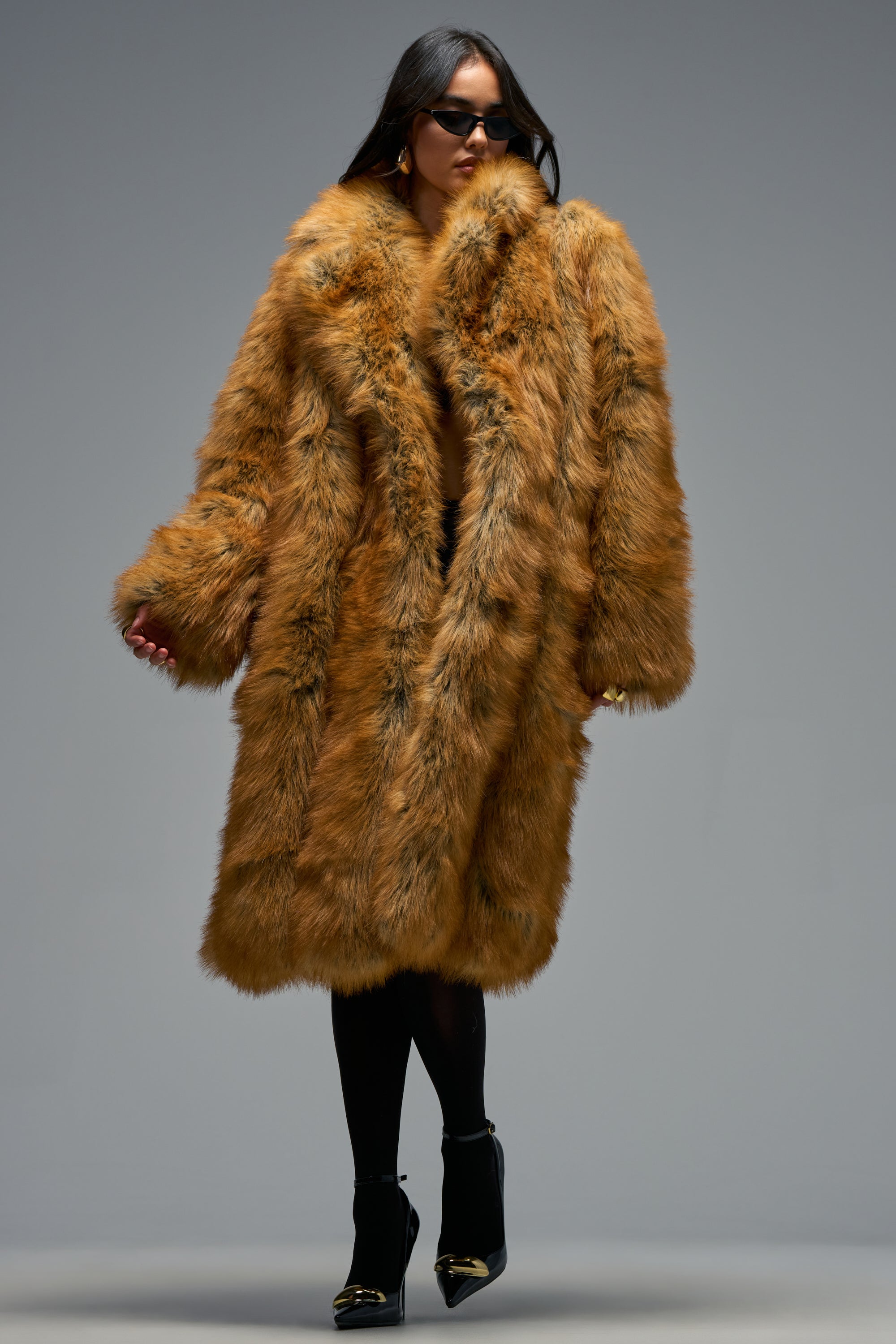 Wearing the BEAUMONT MAXI FAUX FUR coat, paired with black leggings, black high-heeled shoes, and sunglasses, a person stands against a plain gray background, effortlessly showcasing elegant winter layering.
