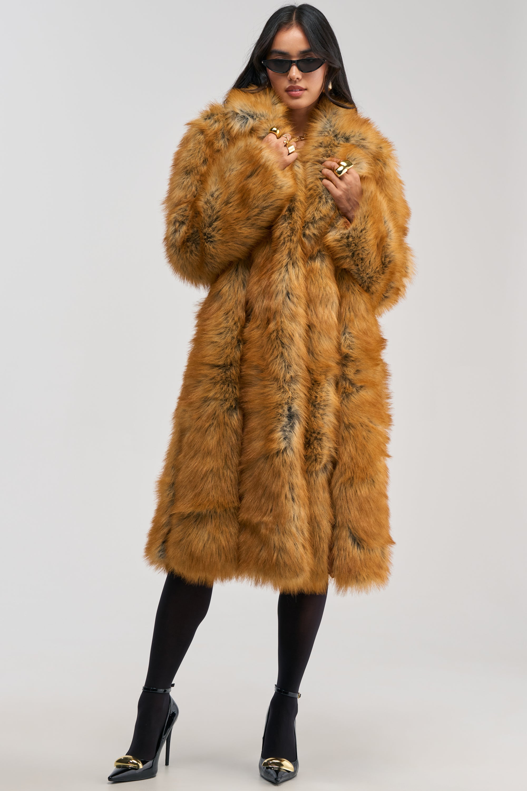 A woman wearing sunglasses, the BEAUMONT MAXI FAUX FUR—a luxurious golden brown longline faux fur coat—styled with black tights and black high heels, stands confidently against a plain light background for an effortlessly chic winter layering look.
