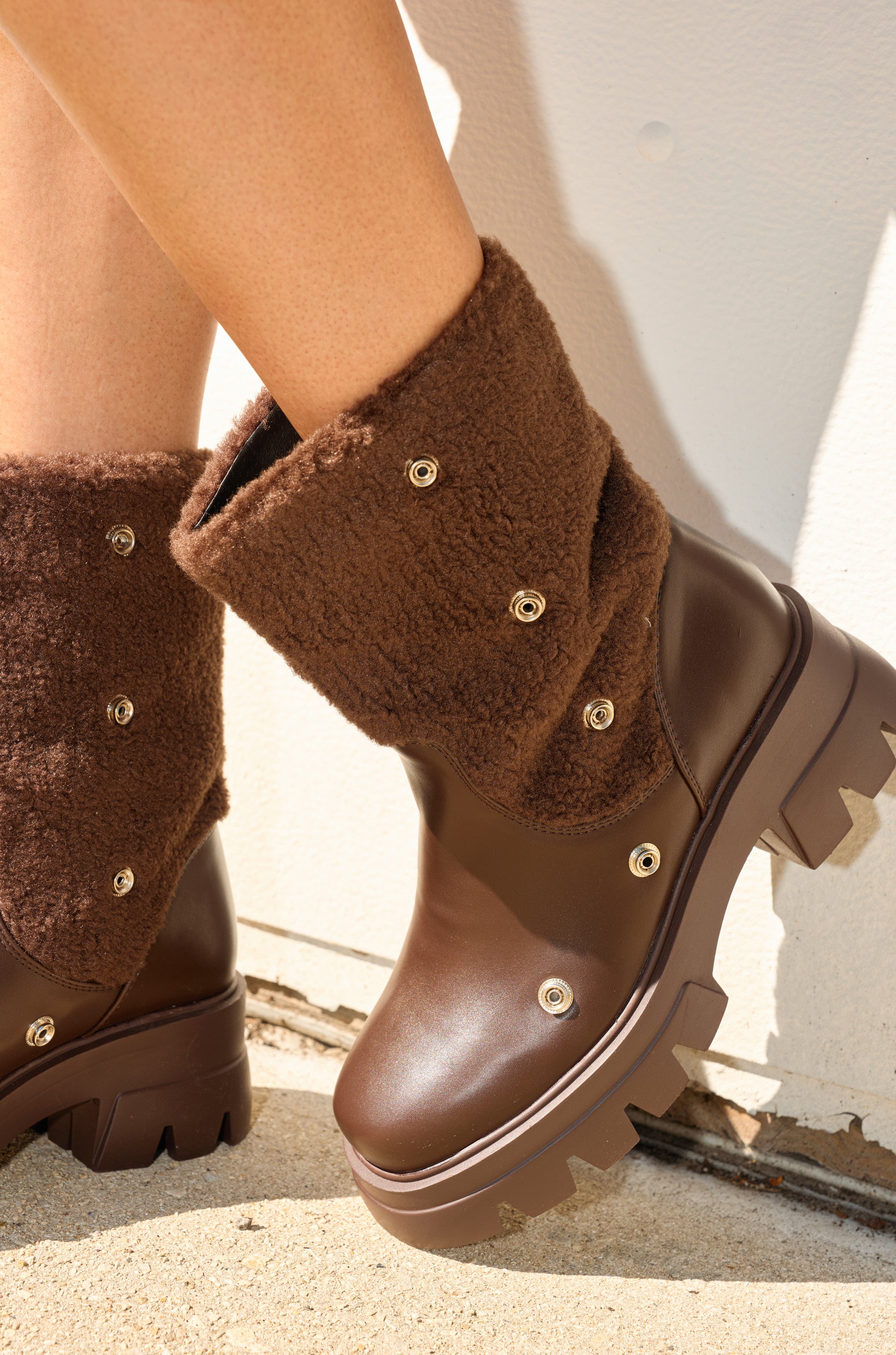 A close-up of a person wearing AZALEA WANG BEHATI BROWN SHERPA BOOTIES with textured, shearling-like cuffs and gold-tone snap details, standing on a light concrete surface near a white wall.