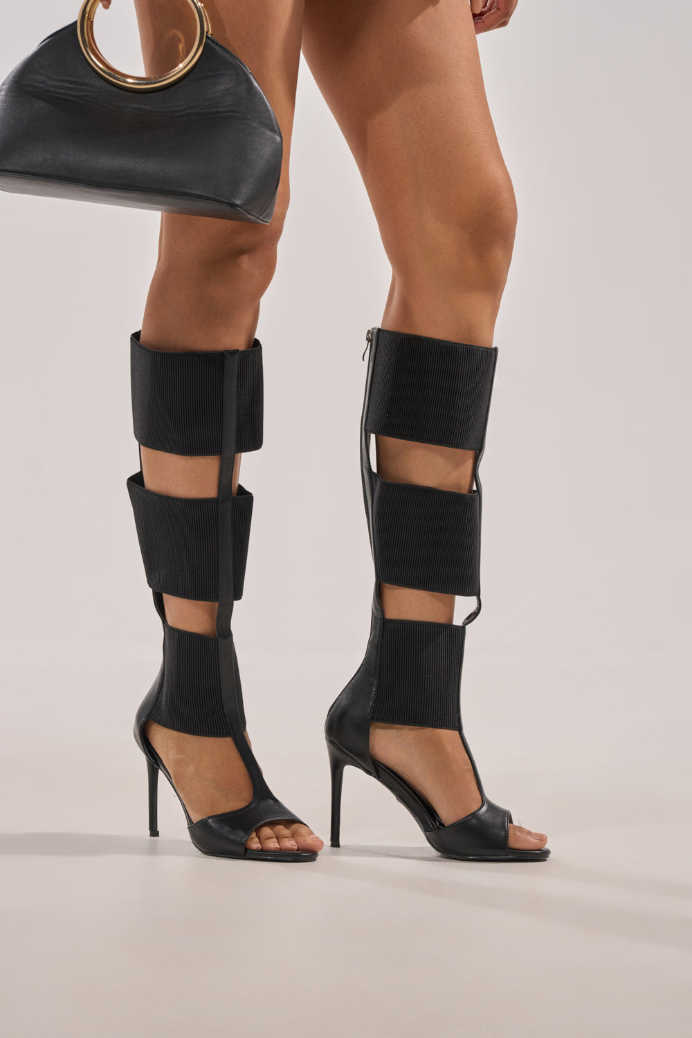 A person wearing AKIRA GOLD BELLANTRA BLACK GLADIATOR STILETTO SANDAL with multiple black straps up to the knees, styled with a black handbag featuring a bold gold circular handle. 2