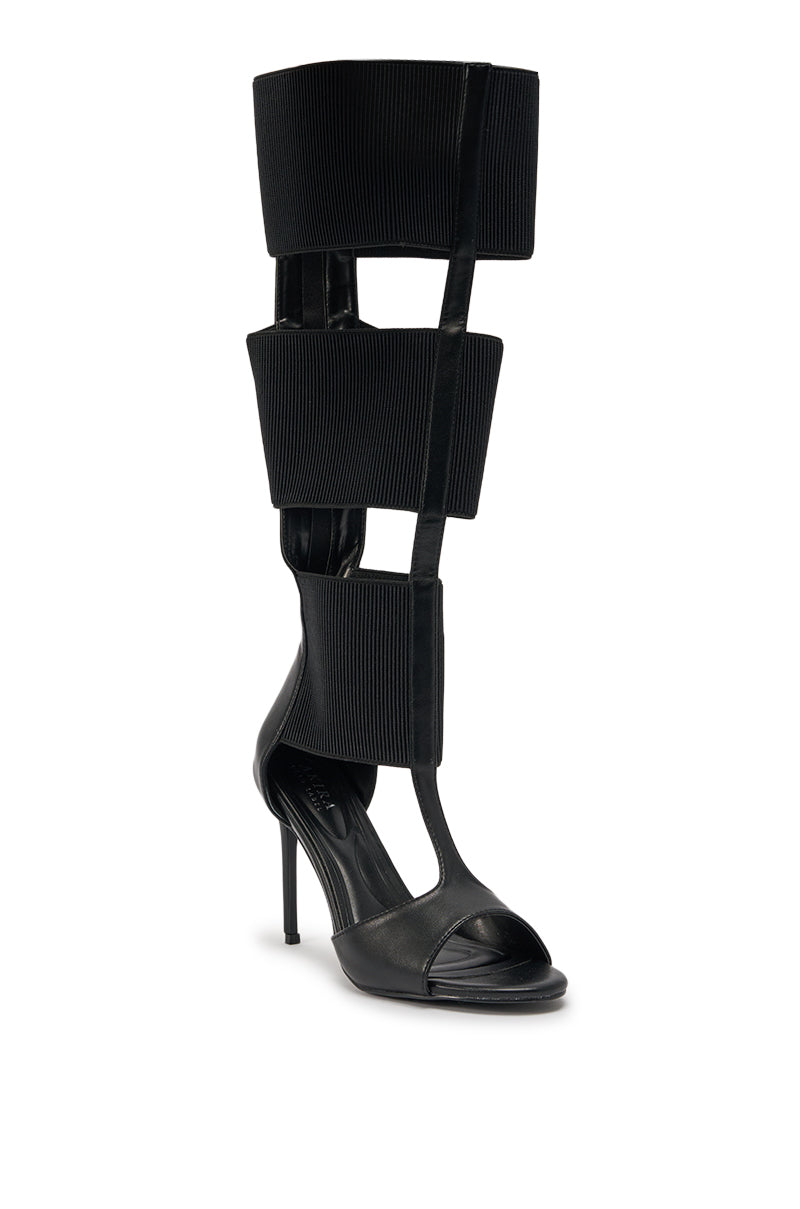 The AKIRA GOLD BELLANTRA BLACK GLADIATOR STILETTO SANDAL is a black faux leather, knee-high gladiator sandal with an open toe, three large rectangular panels joined by vertical straps, and a sleek stiletto heel. 4