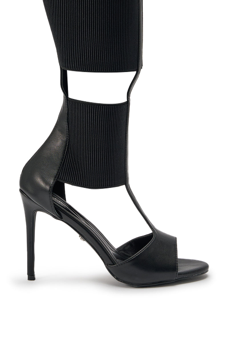 The AKIRA GOLD BELLANTRA BLACK GLADIATOR STILETTO SANDAL features wide ribbed elastic straps up the leg, an open toe and ankle, and a bold stiletto heel, styled here with black pants for a modern look. 6