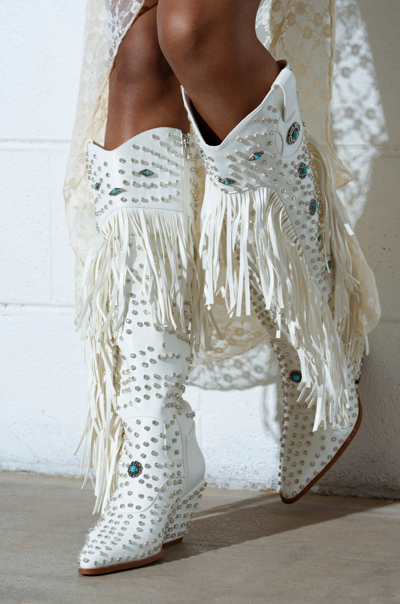 A person wears AZALEA WANG BENNETT WHITE BOOT—knee-high, faux leather boots with studs and turquoise details. Featuring a pointed toe and mid-height heel, the boots pair elegantly with lace attire against a concrete backdrop.