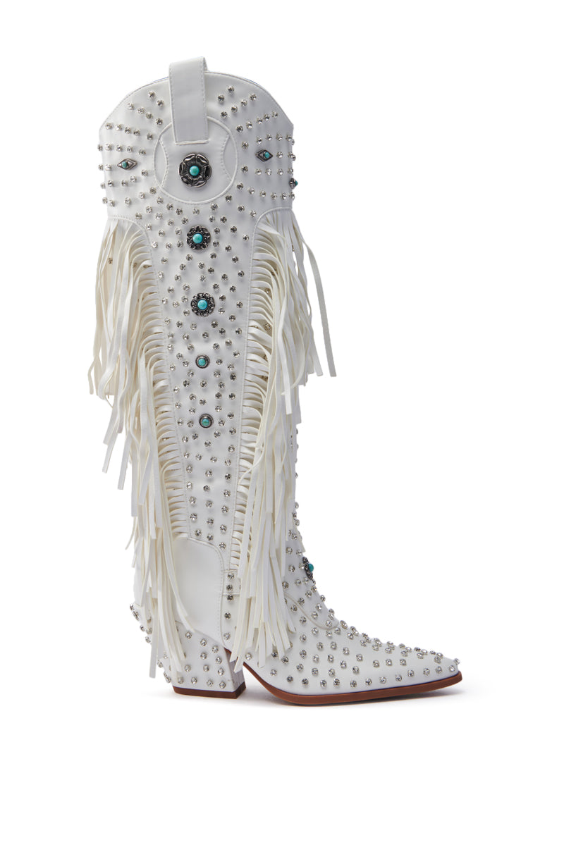 The AZALEA WANG BENNETT WHITE BOOT is a knee-high, faux leather cowgirl boot. It features silver studs, turquoise accents, long white fringe along the sides, a pointed toe, and a decorative circular detail near the top.