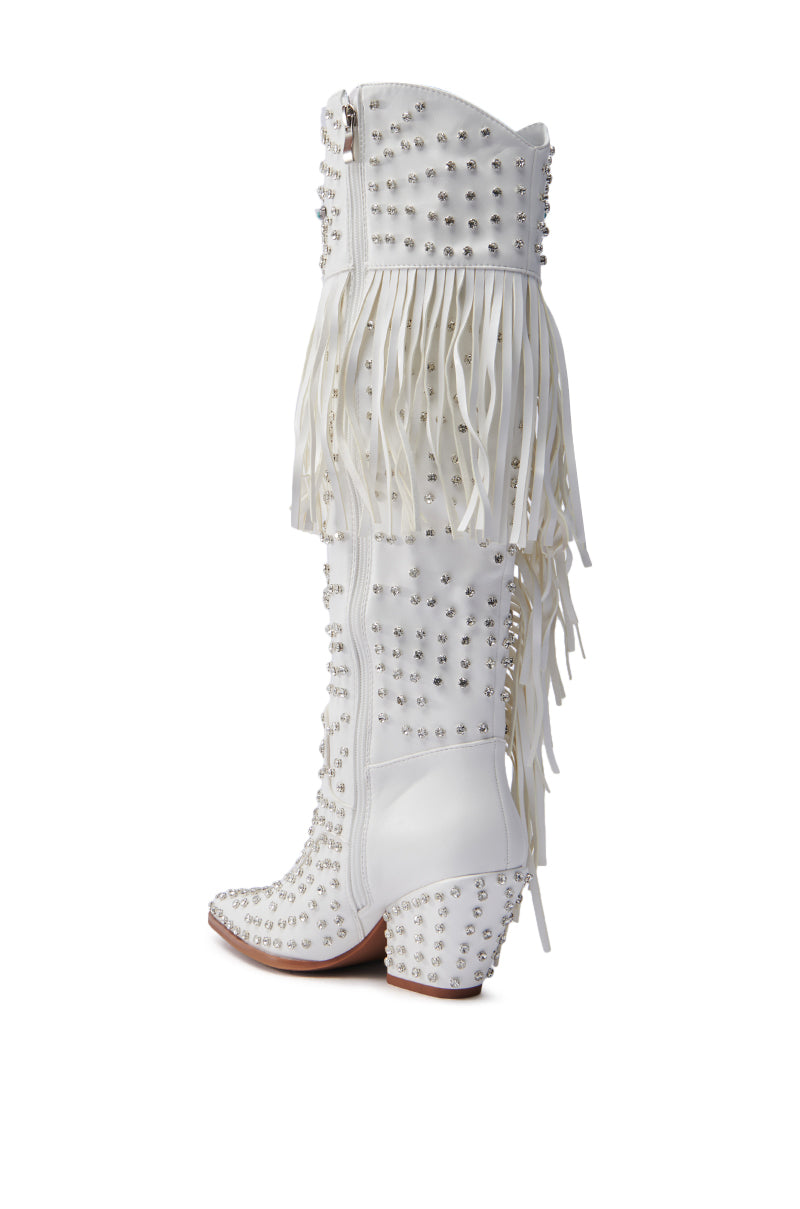 The AZALEA WANG BENNETT WHITE BOOT is a faux leather knee-high cowgirl boot, embellished with silver studs and fringe on the upper section, featuring a mid-high chunky heel and slightly pointed toe for a stylish look.
