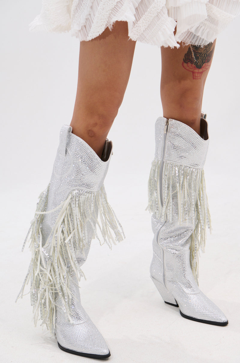 A person in the AZALEA WANG BENNETTO SILVER RHINESTONE WESTERN BOOT, featuring knee-high silver rhinestones and fringe, stands on a white background with a white pleated skirt and a colorful thigh tattoo partially visible.