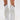 Wearing the AZALEA WANG BENNETTO SILVER RHINESTONE WESTERN BOOT with a snakeskin texture and fringe, paired with a white ruffled skirt and showing off a thigh tattoo, they pose against a plain white background.