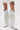 Wearing the AZALEA WANG BENNETTO SILVER RHINESTONE WESTERN BOOT with a snakeskin texture and fringe, paired with a white ruffled skirt and showing off a thigh tattoo, they pose against a plain white background.