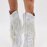 Wearing the AZALEA WANG BENNETTO SILVER RHINESTONE WESTERN BOOT with a snakeskin texture and fringe, paired with a white ruffled skirt and showing off a thigh tattoo, they pose against a plain white background.