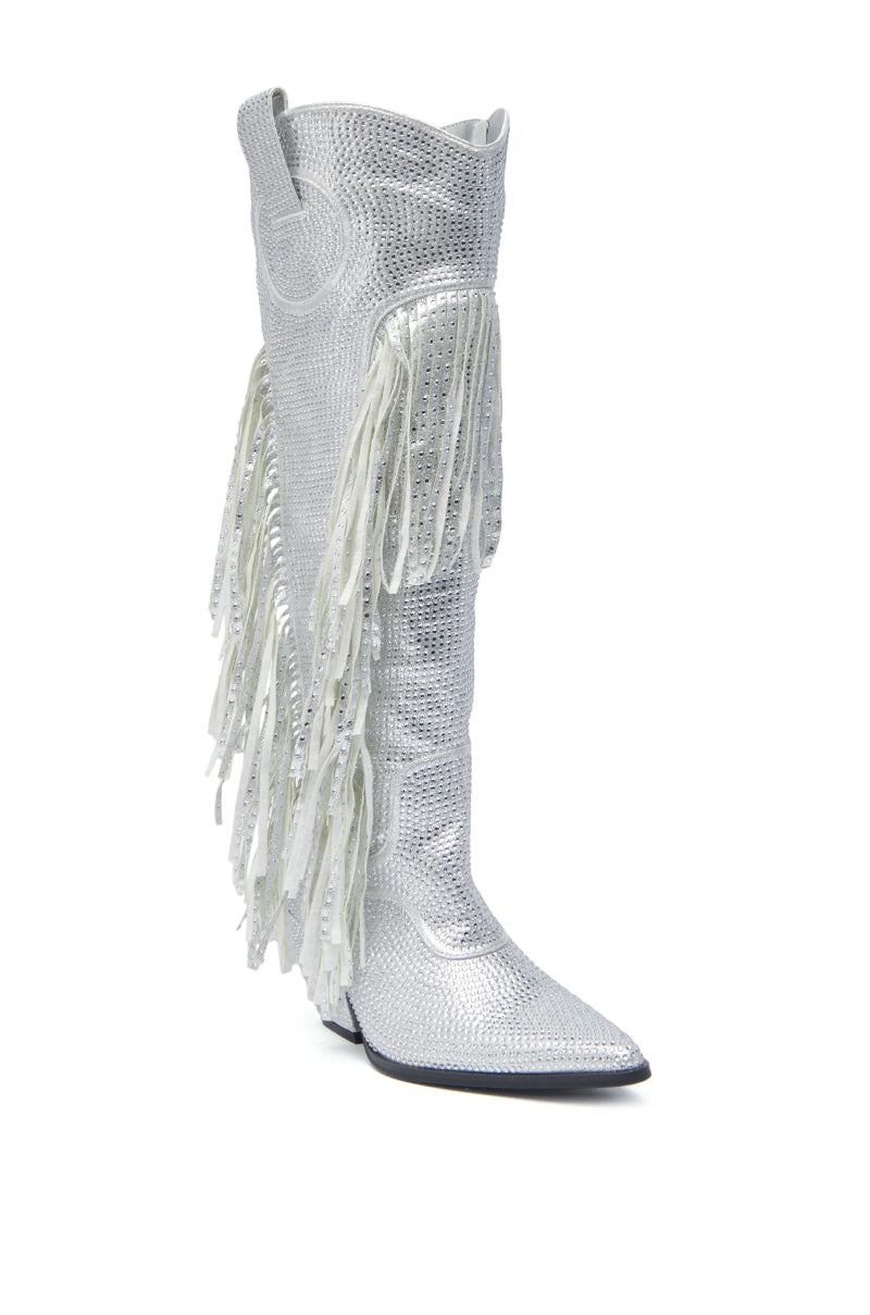 The AZALEA WANG BENNETTO SILVER RHINESTONE WESTERN BOOT is a knee-high, pointed-toe silver boot with rhinestones, a chunky heel, textured surface, and white fringe along the sides, shown on a white background.