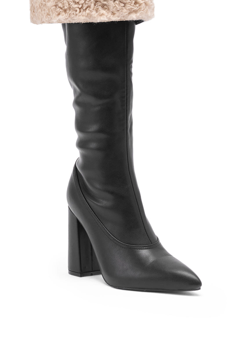 A person wears the AZALEA WANG BENOUL BLACK SHERPA LINED CHUNKY BOOT, featuring a high block heel and pointed toe, styled with beige faux fur or shearling at the top for a cozy-chic look.