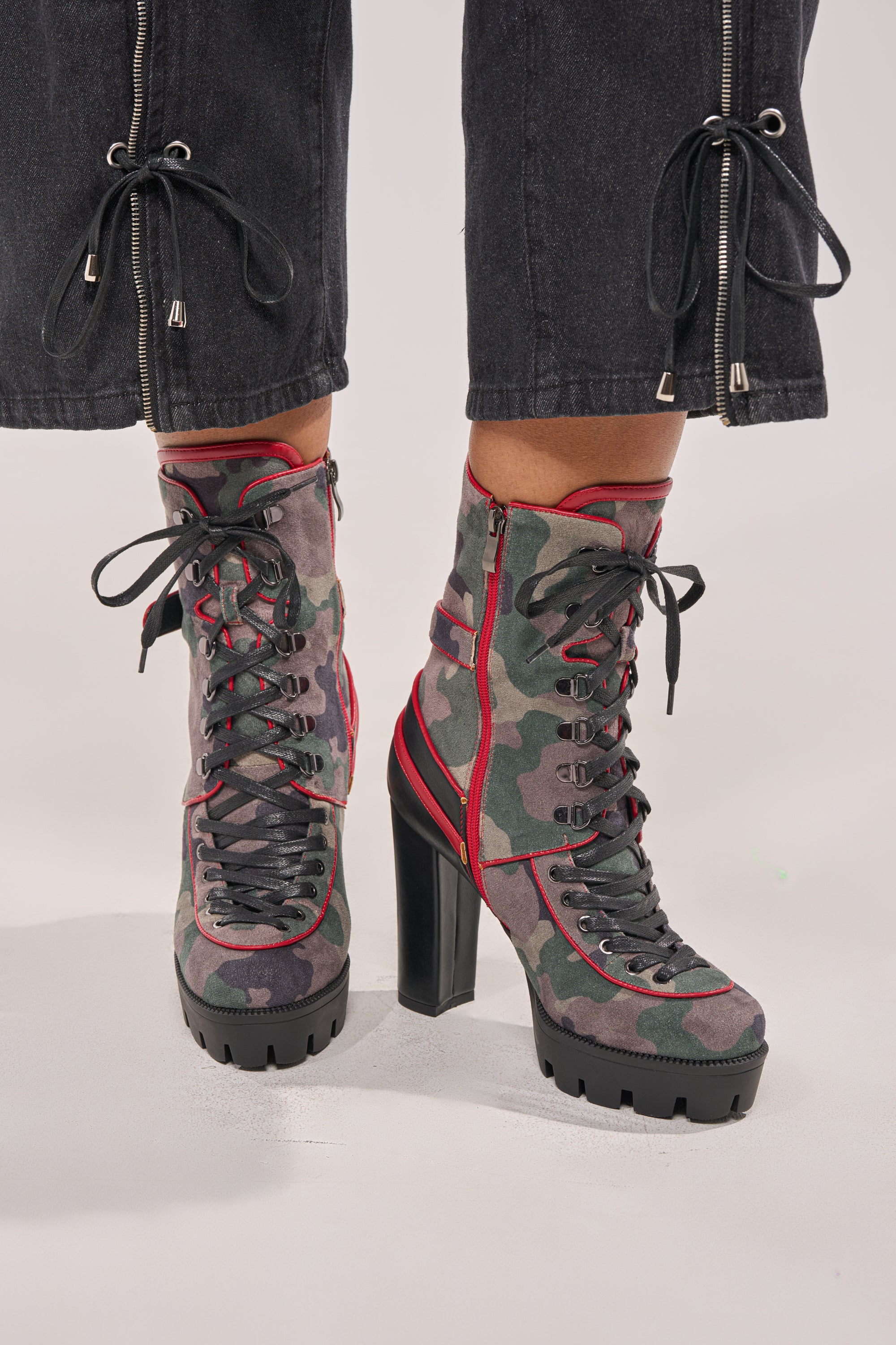 A person in cropped black zipper pants wears the AZALEA WANG BENSON CAMO BLOCK SPORTY BOOTIE—high-heeled platform booties with a green camo pattern, chunky block heels, lug sole, and bold red trim for standout streetwear style. 0