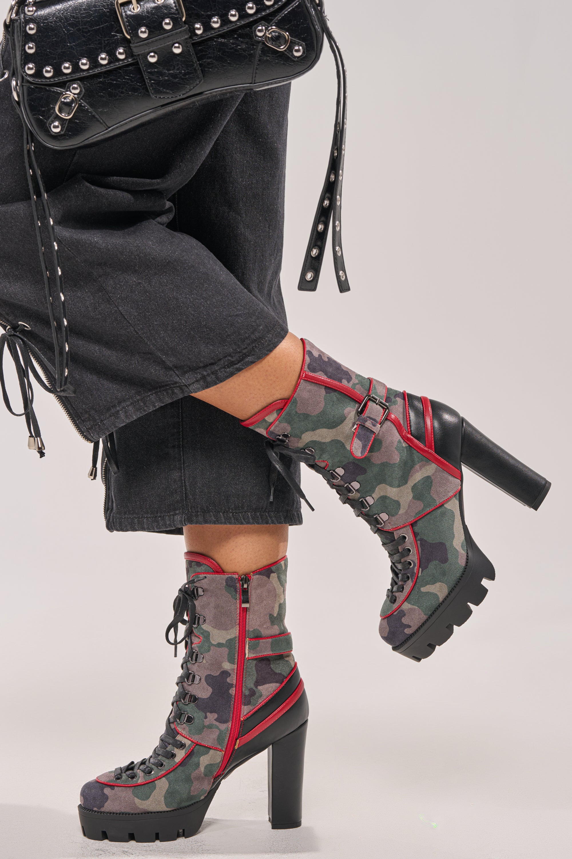Wearing black wide-leg pants, a person rocks the AZALEA WANG BENSON CAMO BLOCK SPORTY BOOTIE—block heel camo booties with red trim and buckles—paired with a black studded handbag and edgy platform lug soles, legs crossed at the ankles. 1