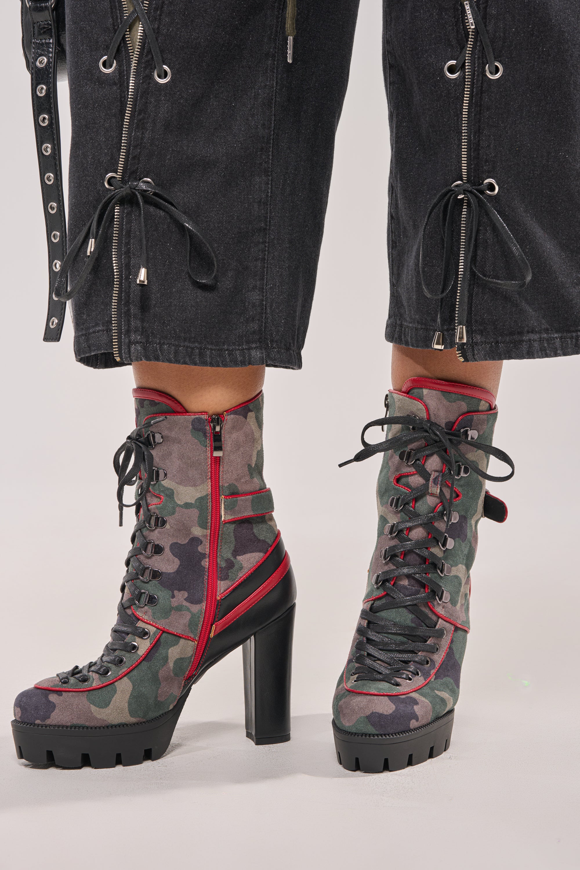 Wearing black jeans with zipper and tie details, a person pairs them with the AZALEA WANG BENSON CAMO BLOCK SPORTY BOOTIE, featuring green and red camo block heels, while standing on a light-colored surface. 2