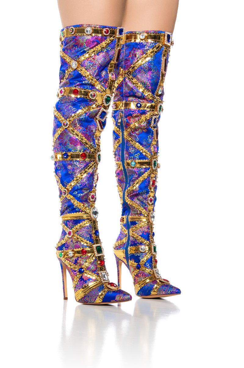 The AZALEA WANG BERKLEY BLUE BROCASE STILETTO BOOT features thigh-high, pointed-toe brocade stiletto boots adorned with a vibrant purple and blue pattern. Gold crisscrossing straps and rhinestone embellishments add a touch of elegance, while the sleek stiletto heels enhance their sophisticated look. Captured against a pristine white background, these boots make a bold fashion statement.