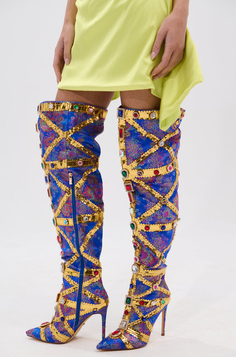 A person in a light yellow skirt and AZALEA WANG BERKLEY BLUE BAROCADE EMBELLISHED BOOTS with colorful gems and stiletto heels stands against a plain white background.