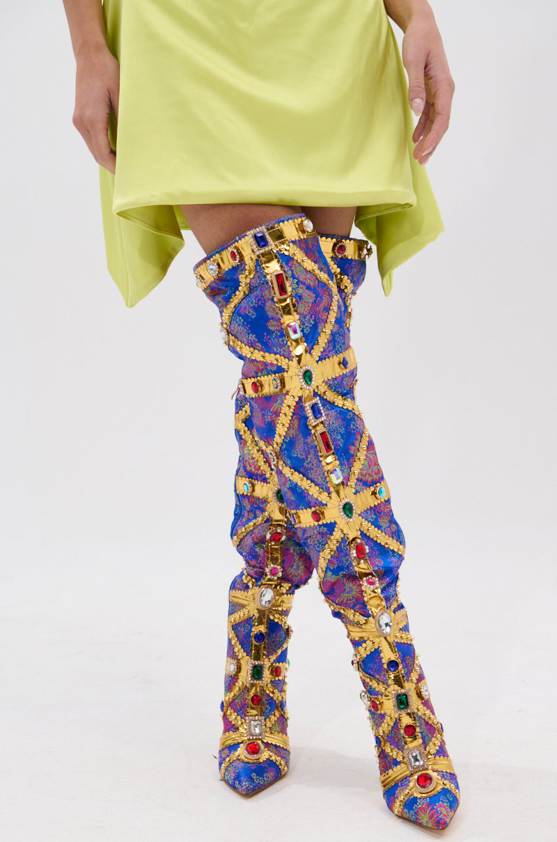 A person wearing a light green skirt and the AZALEA WANG BERKLEY BLUE BAROCADE EMBELLISHED BOOT, featuring gold straps and multicolored gemstones, stands on a white background.
