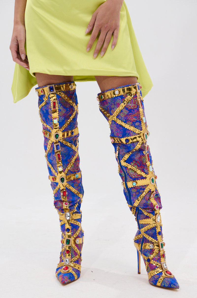 A person stands on a white background wearing a light green skirt and AZALEA WANG BERKLEY BLUE BROCADE EMBELLISHED BOOTS—thigh-high stilettos adorned with colorful jewels and gold detailing.