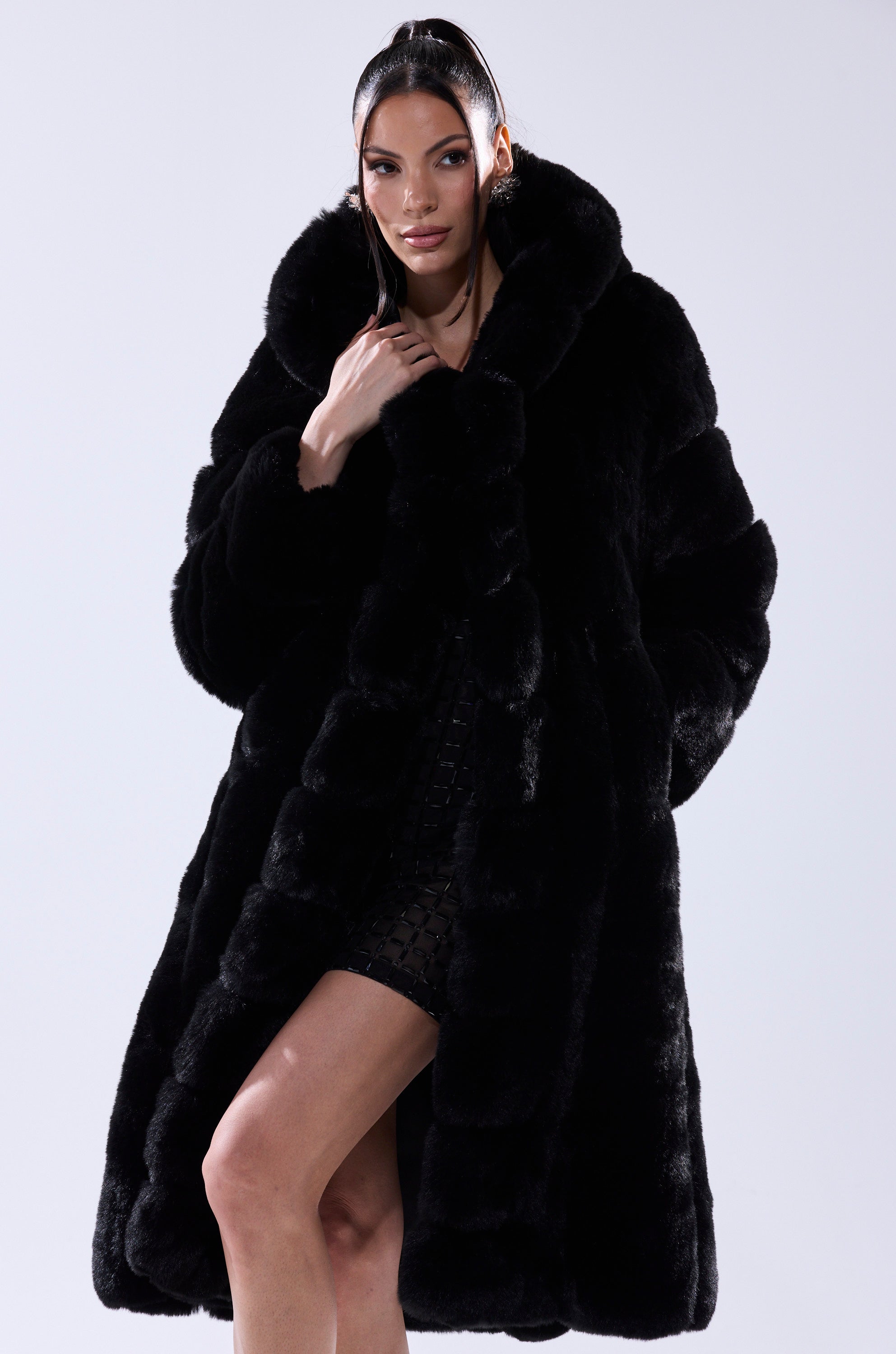 A woman poses confidently in the BERLIN HOODED FAUX FUR IN BLACK, its longline silhouette draped over a short black dress. She stands against a plain light background, her hand gently touching the coat's collar.