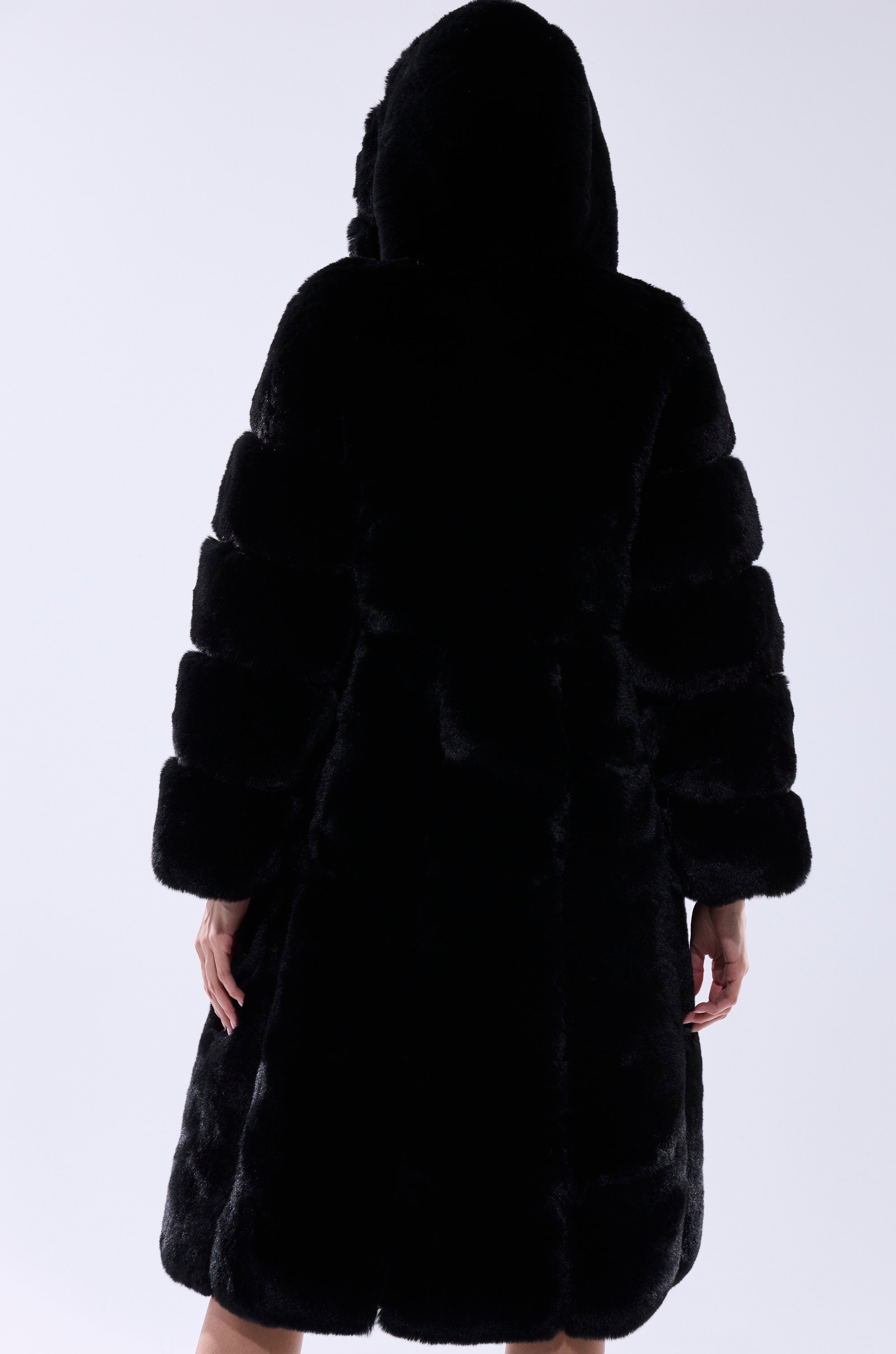 A person faces away, wearing the BERLIN HOODED FAUX FUR IN BLACK. This black longline jacket has a hood, horizontal sections on the sleeves, and falls just above the knees, set against a plain white background.