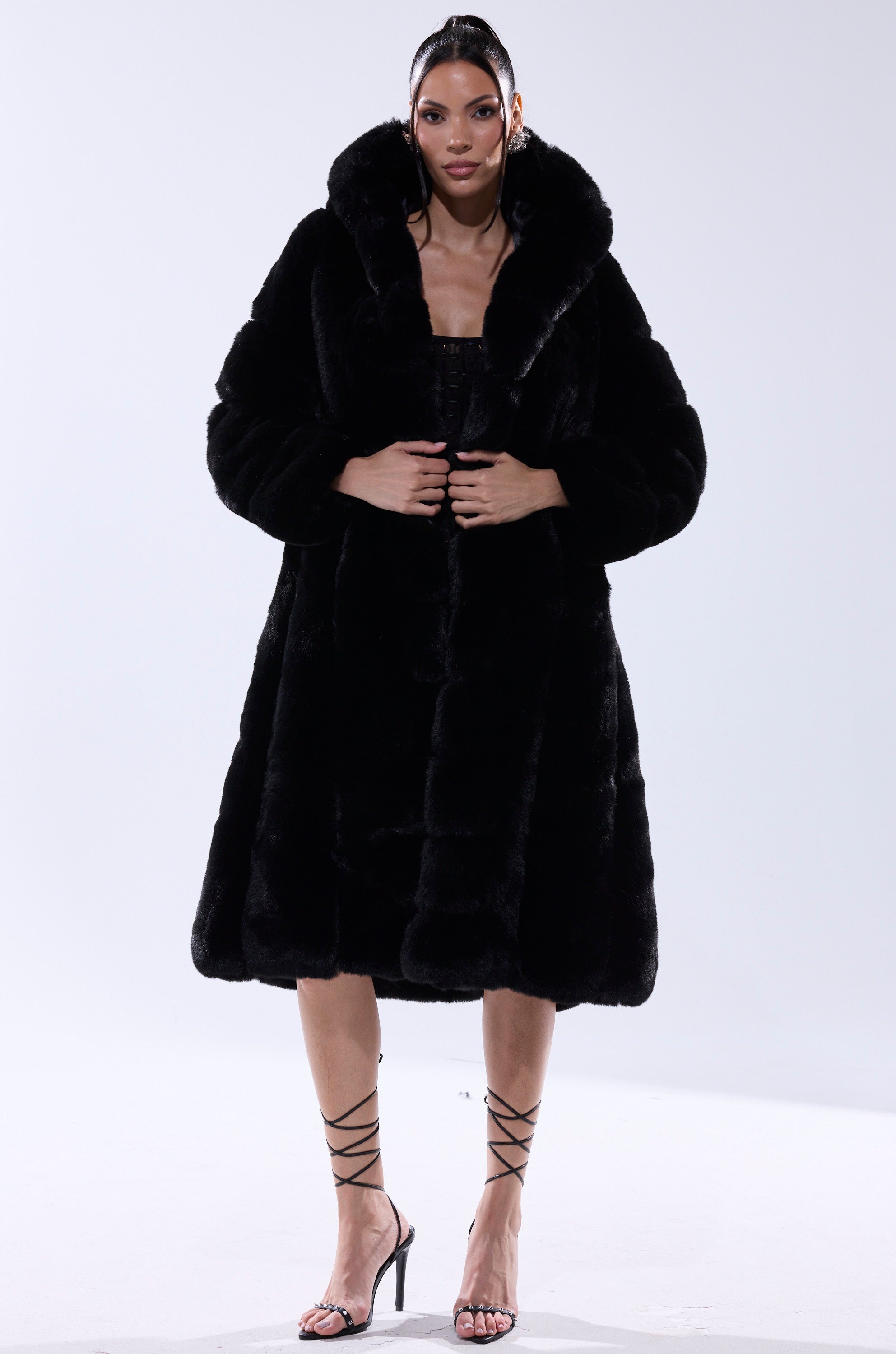 A woman stands against a plain background, wearing the BERLIN HOODED FAUX FUR IN BLACK with a large collar, strappy black heels, and her hair pulled back.