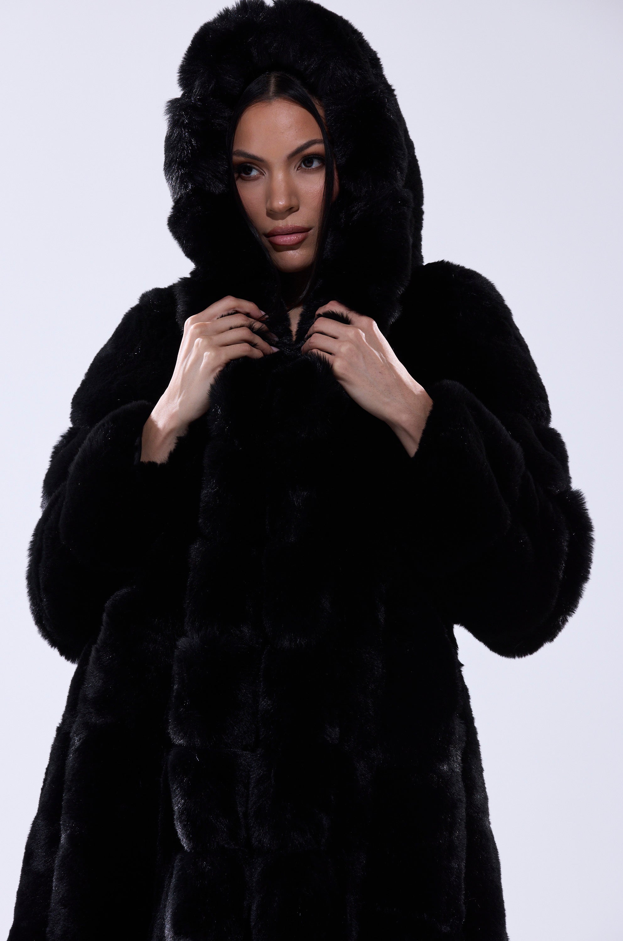 A woman wears the BERLIN HOODED FAUX FUR IN BLACK, featuring a longline silhouette and hood, as she holds the hood with both hands and looks forward against a plain light background.