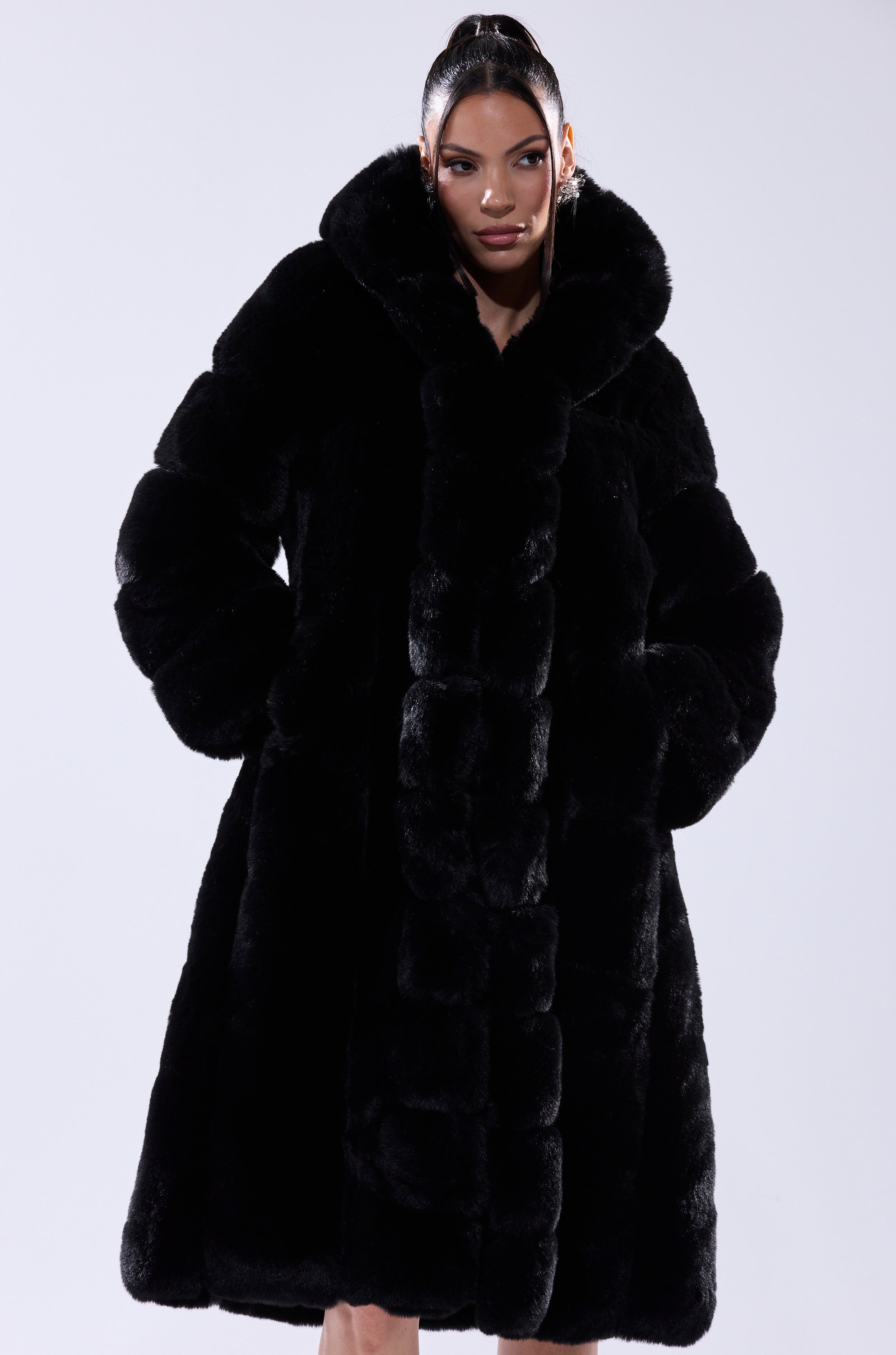 A woman stands against a plain background, hands in pockets, wearing the BERLIN HOODED FAUX FUR IN BLACK, and looks confidently at the camera.