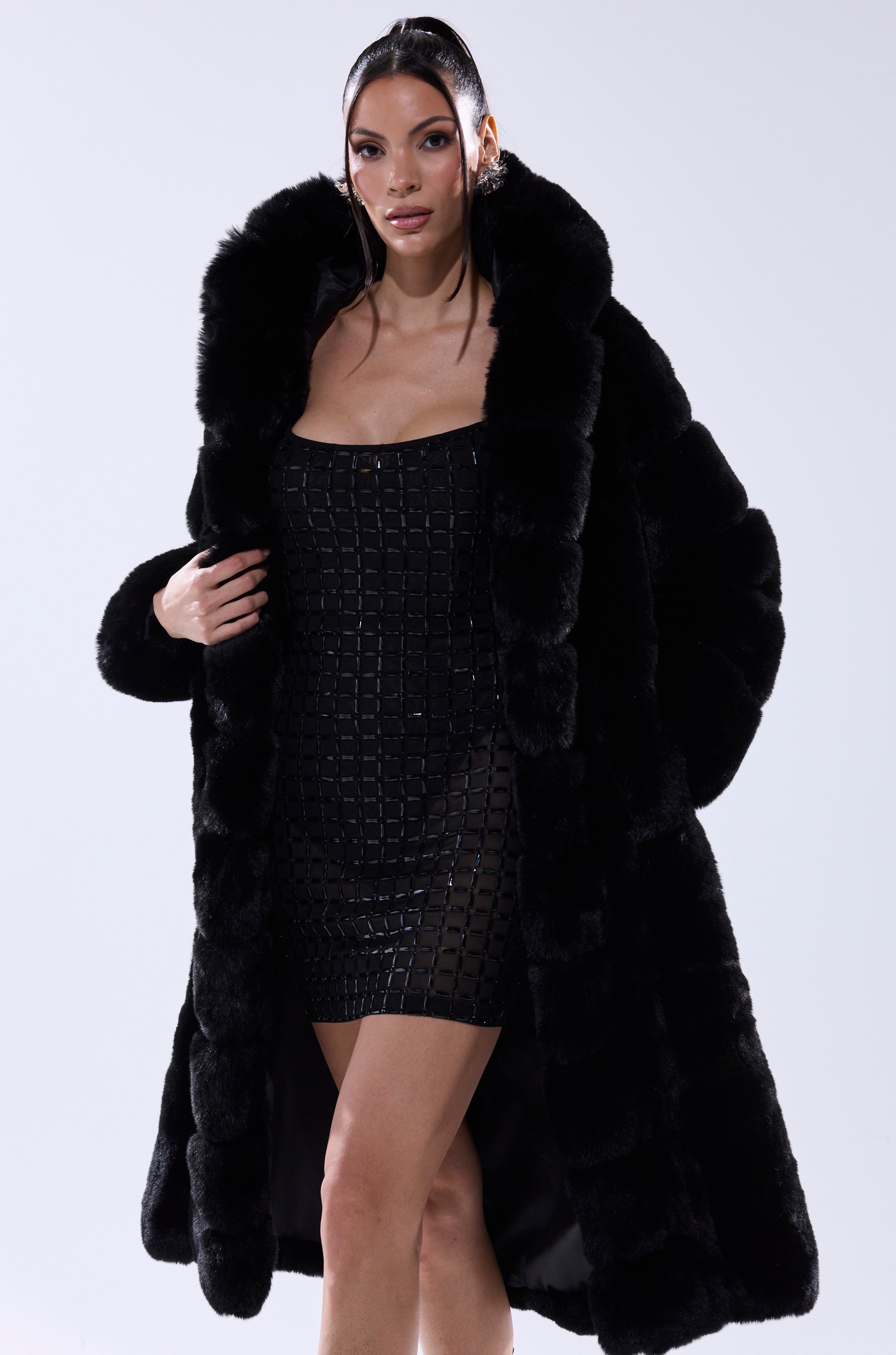 A woman poses in the BERLIN HOODED FAUX FUR IN BLACK, a luxurious longline jacket, over a strapless grid-patterned black and silver mini dress against a plain white background.