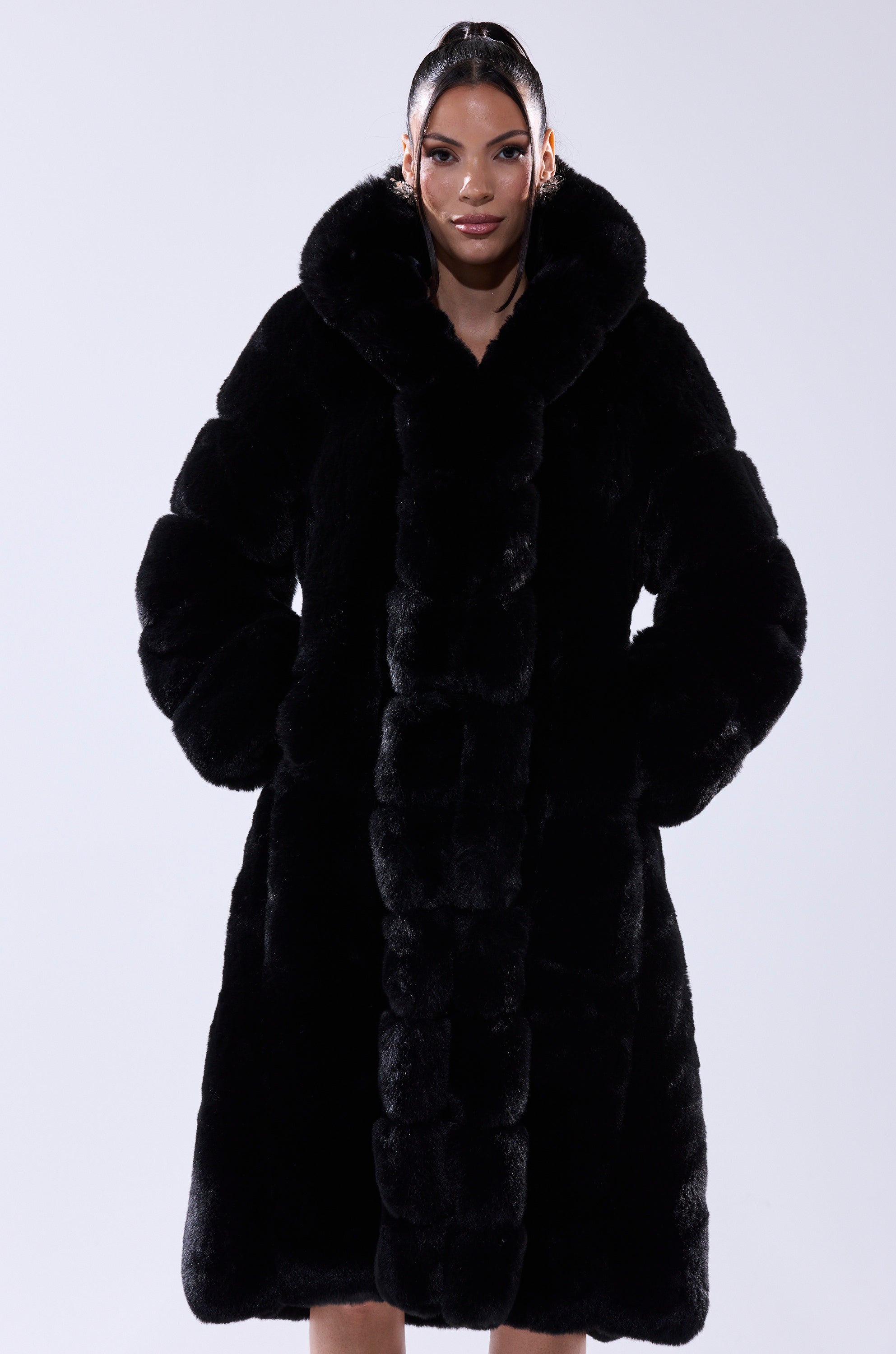 A woman stands facing forward, wearing the BERLIN HOODED FAUX FUR IN BLACK with a large hood and her hands in the pockets against a plain light background.
